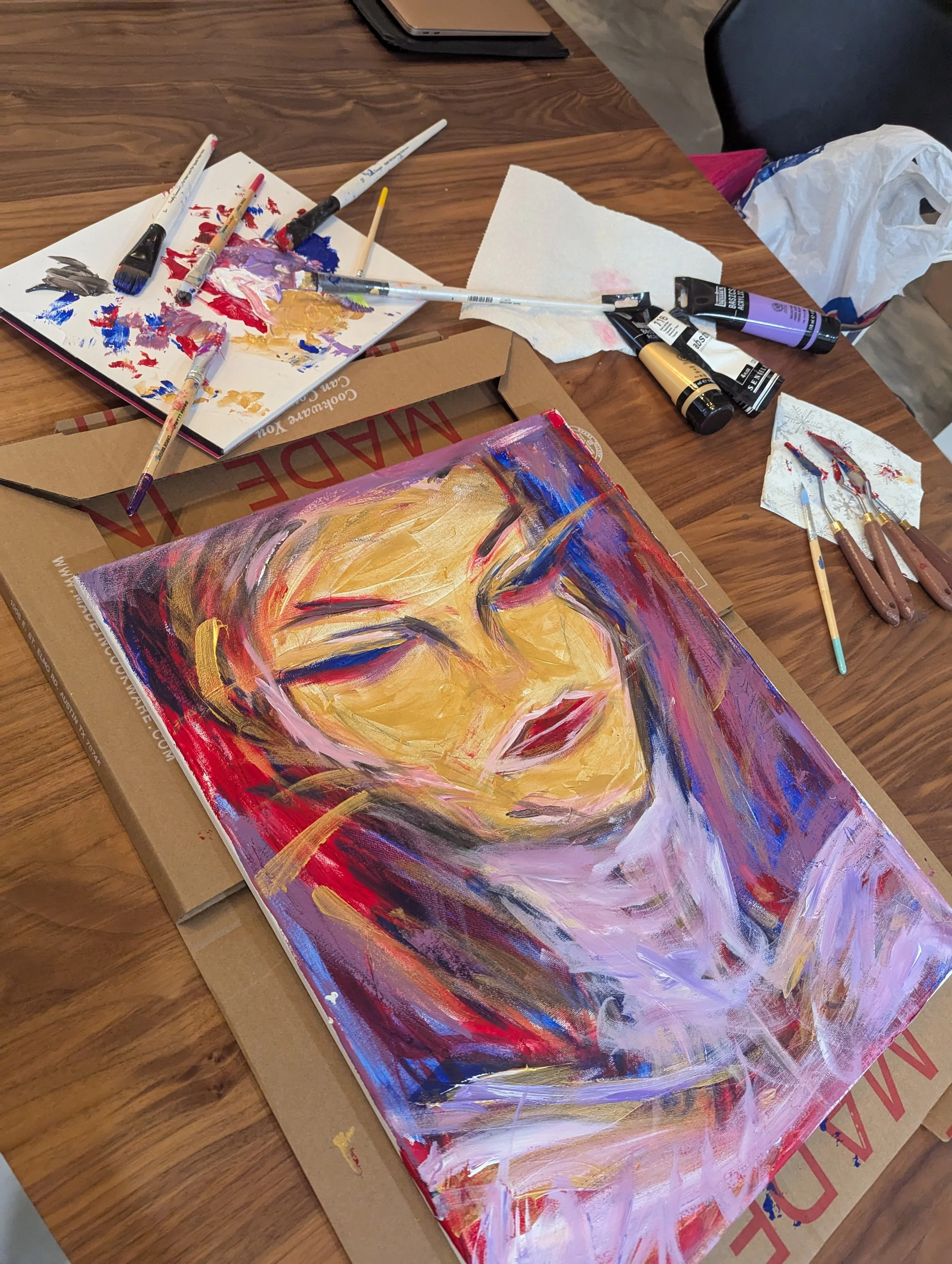 An abstract portrait painting of a person's face with closed eyes, using vibrant colors and bold brushstrokes, on a canvas lying on a table with painting supplies around.