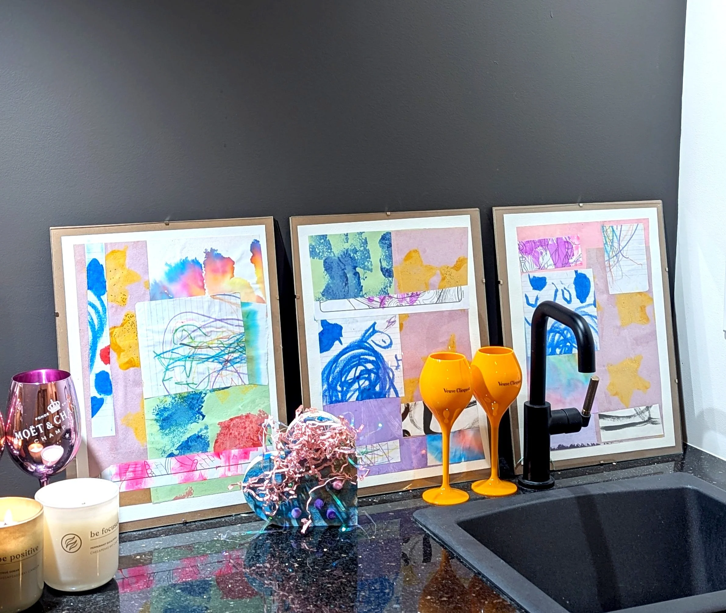 Decorative kitchen countertop with three framed abstract art pieces, two orange champagne glasses, a black faucet, a pink Moët & Chandon glass, two candles, and a decorative item with colorful ribbons.