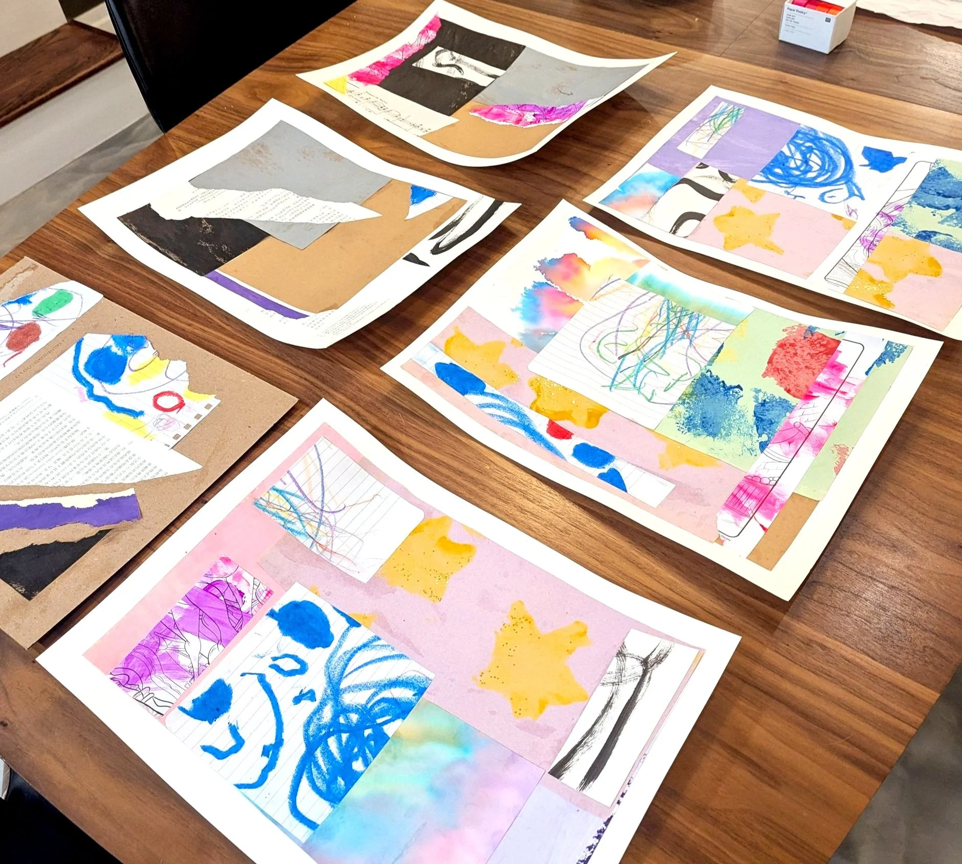 Colorful abstract art collages and sketches on paper, spread out on a wooden table.