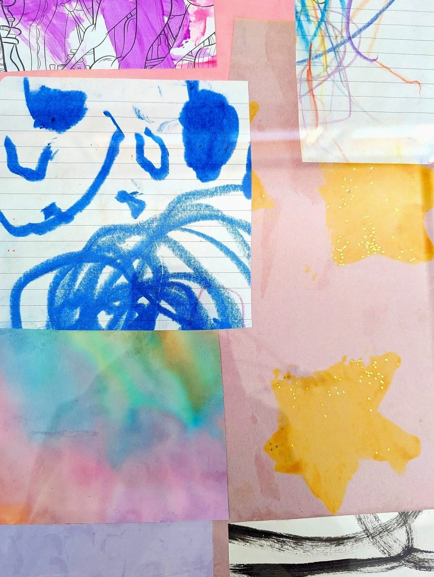 Child's artwork on lined paper featuring a blue crayon drawing of a smiling face with two eyes, a nose, a mouth, and a curled shape below, along with various colorful paper and crayon scribbles, paint splashes, and glitter on a pink background.