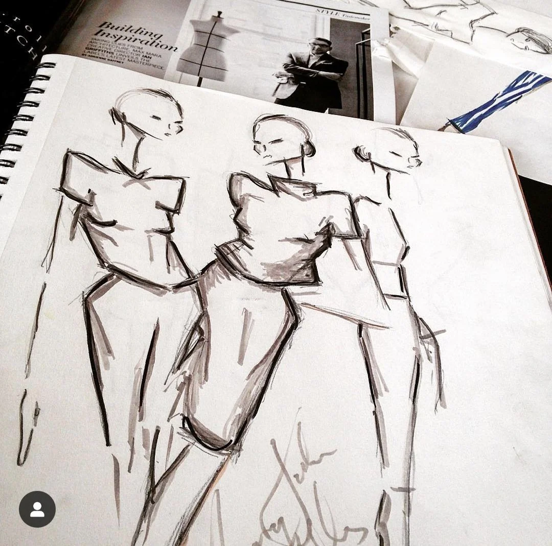 Sketch of three women in fashionable clothing, drawn with black ink on a sketchbook. The sketch shows women in stylish outfits with confident poses. Papers with photographs and text are in the background.