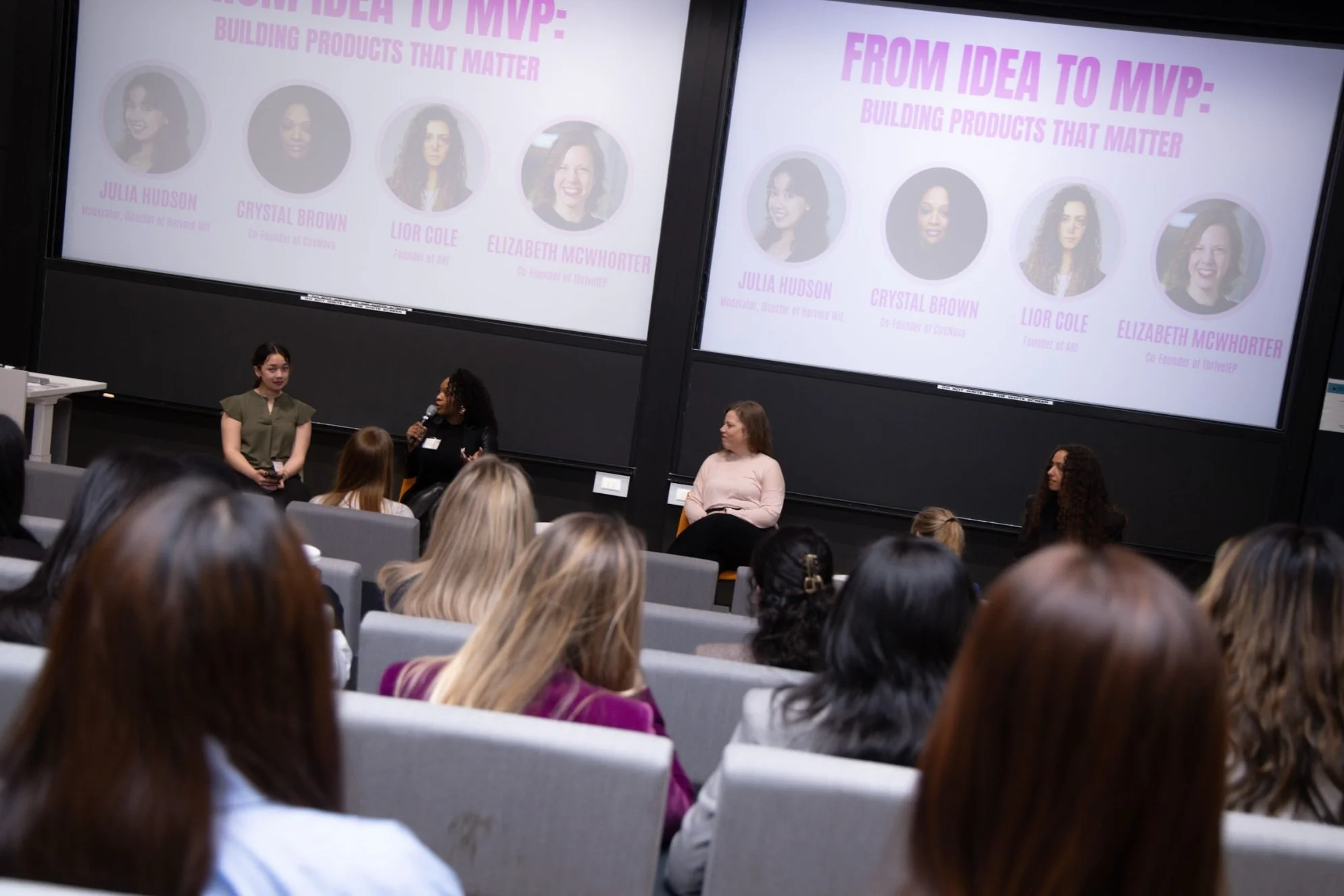 Women speaking and sitting on stage in front of a large screen, during a conference or panel discussion, with an audience seated and listening.