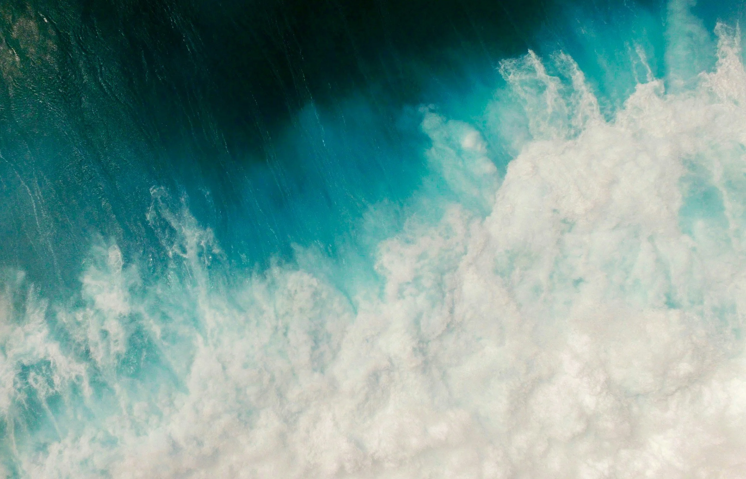 Aerial view of a large ocean wave with white foam at the crest, blue water, and a hint of green.