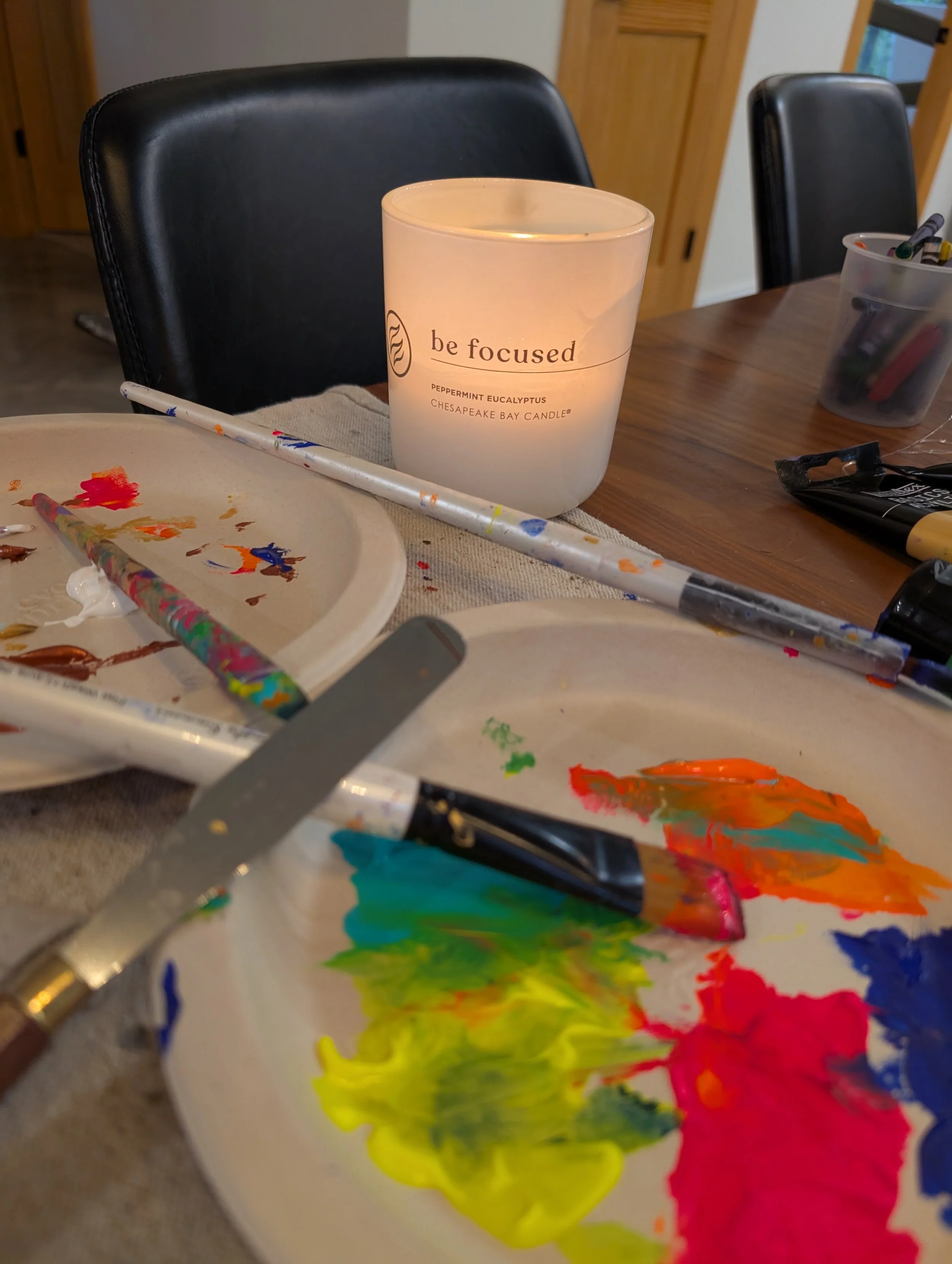 Painting supplies on a table, including a palette with multicolored paint, paintbrushes, and a lit candle with the words 'be focused' printed on it.