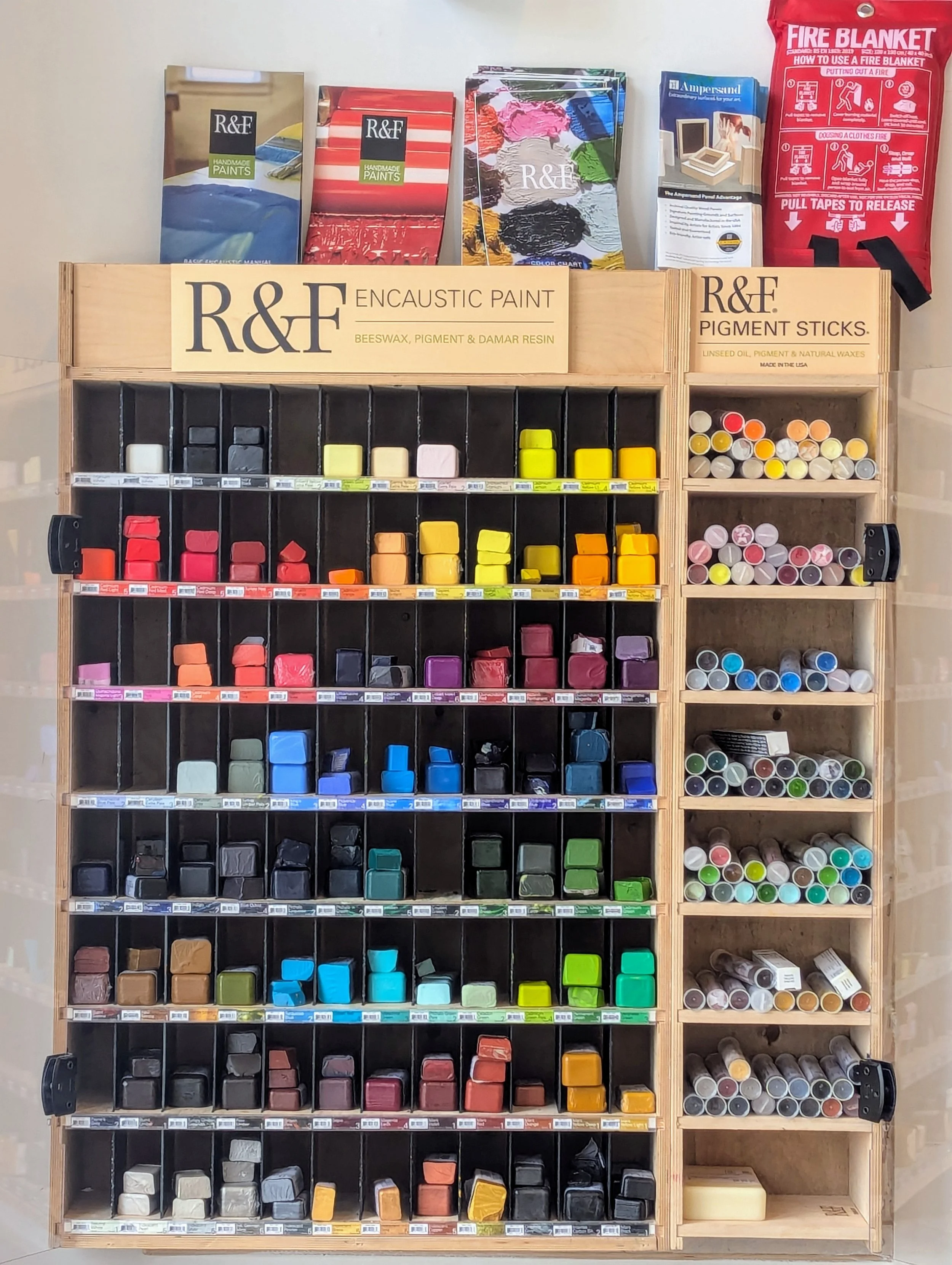Display of R&F encaustic paint, pigment sticks, and related painting supplies organized on a wooden shelf with multiple color options.