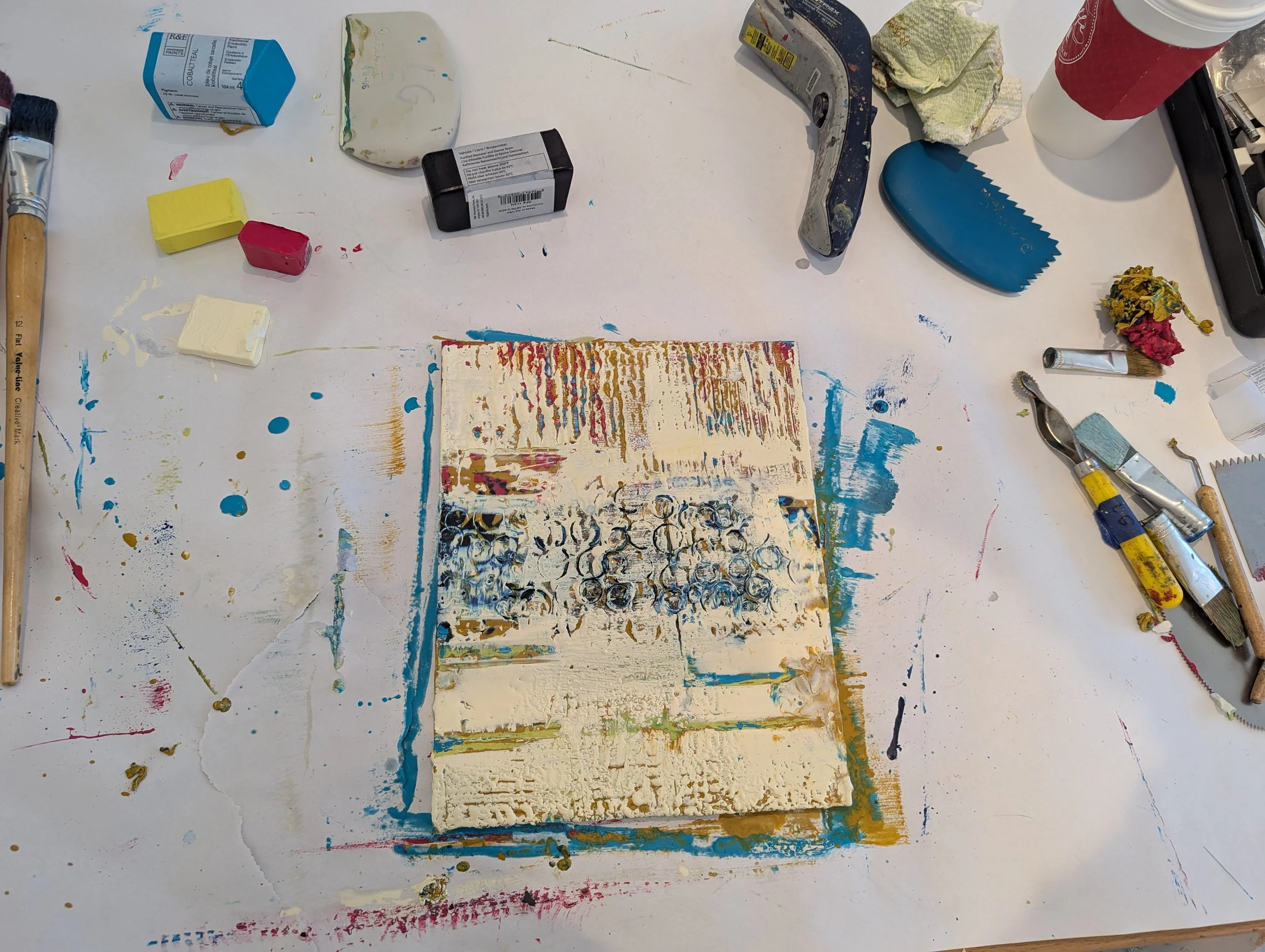 A cluttered artist's workspace with tools, paints, and a textured canvas or paper at the center, spread out on a white table with paint splatters and spots.