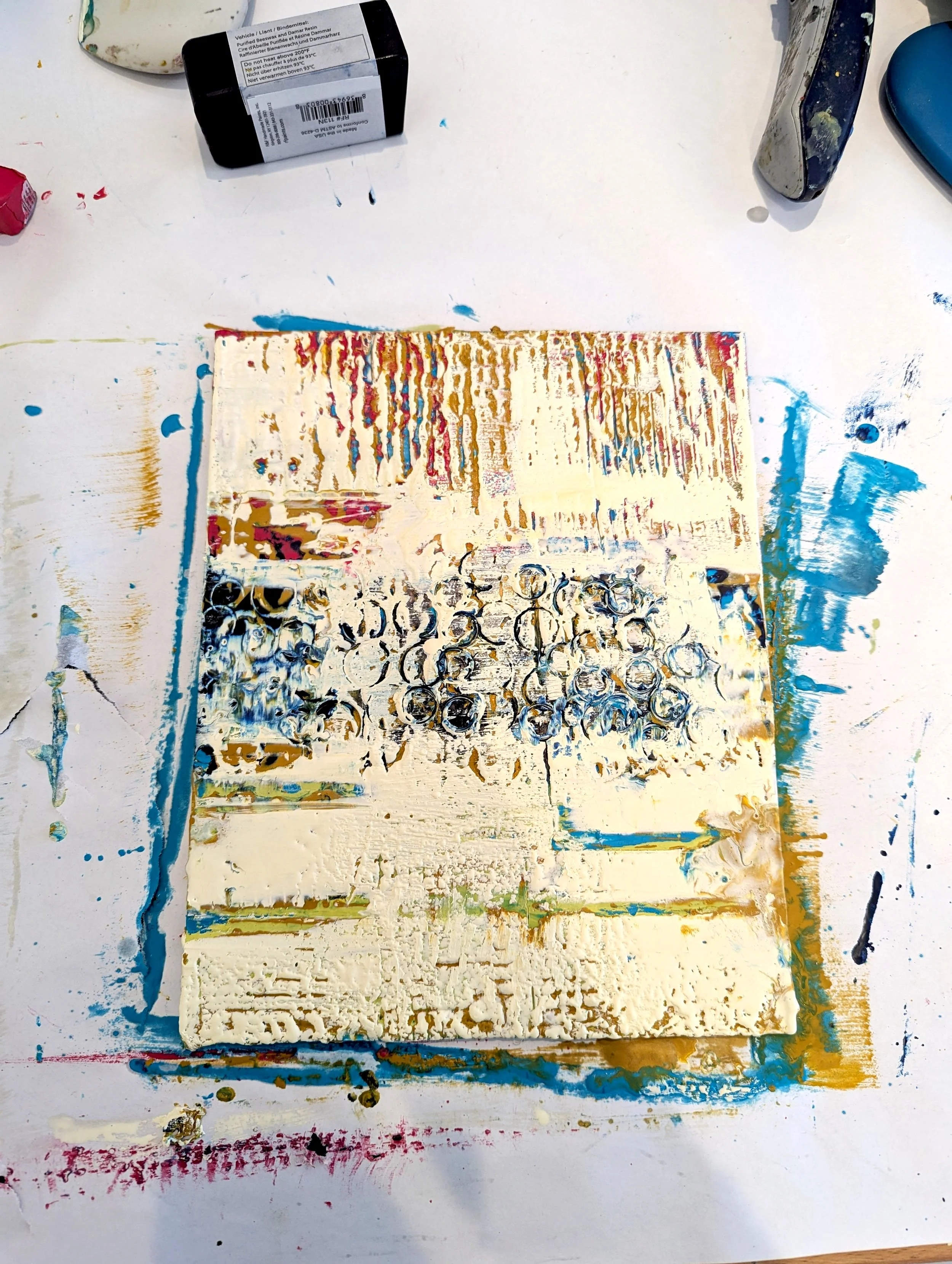 Abstract mixed media artwork with textured layers of cream, blue, red, and gold colors, featuring circular patterns and drips, on a white background surrounded by paint splatters and tools.