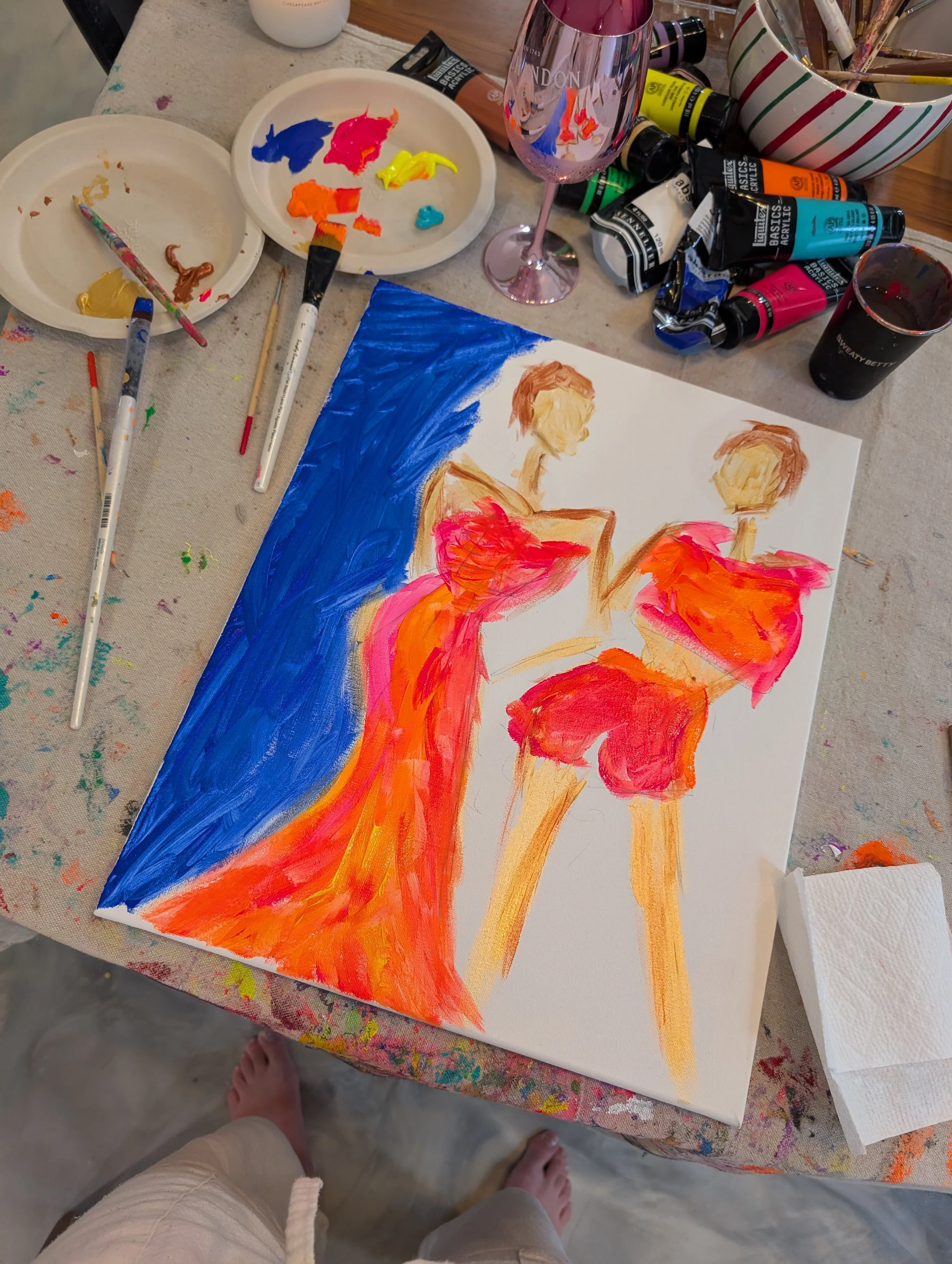 A painting of two women wearing vibrant red dresses in progress, with painting supplies and brushes scattered on a table covered with paint stains.