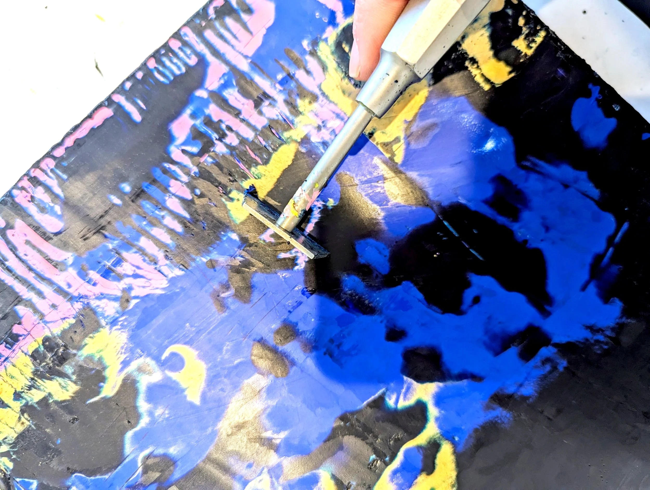 Close-up of a person using a spray gun to create abstract spray paint art on a black canvas, with prominent blue, yellow, pink, and purple colors.