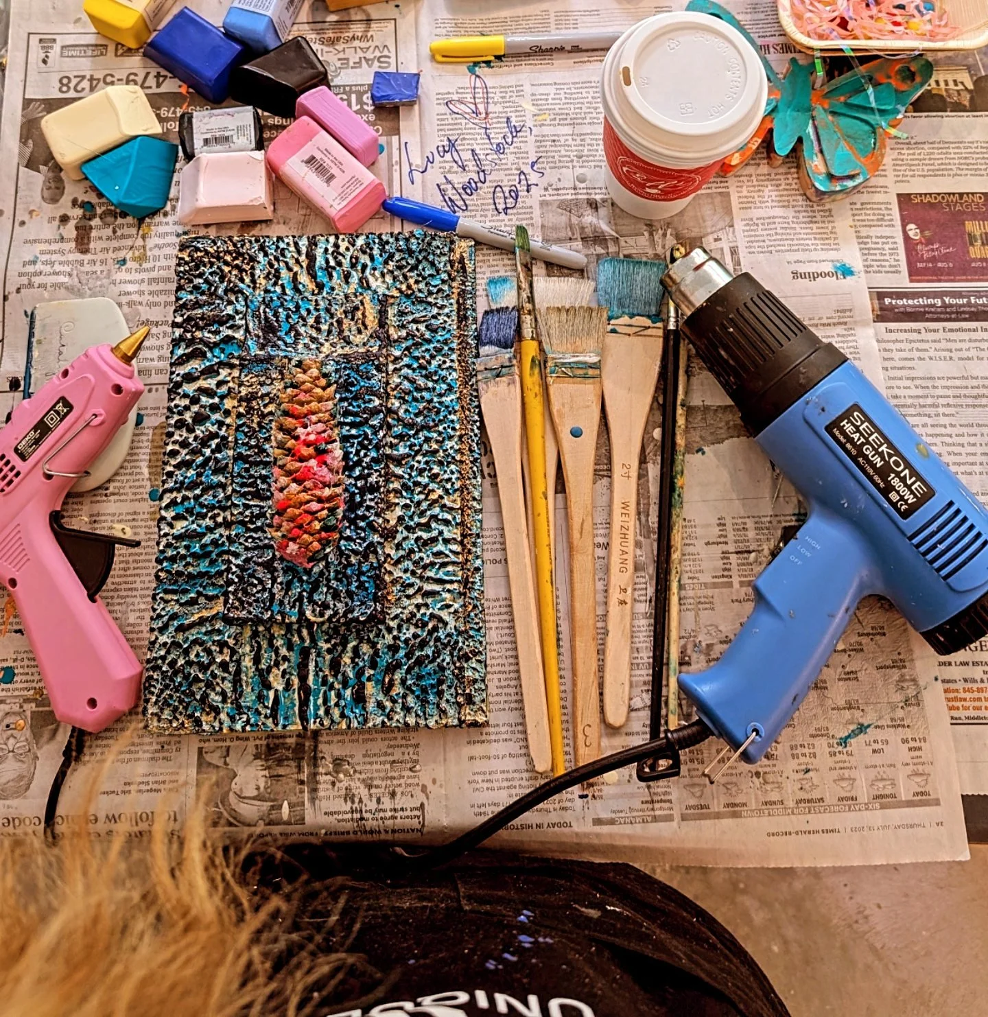 Art supplies on newspaper-covered surface including hot glue gun, mini glue sticks, paint brushes, bottles of paint in pastel colors, a cup of coffee, and a colorful butterfly decoration. A small textured canvas with abstract painting is also present