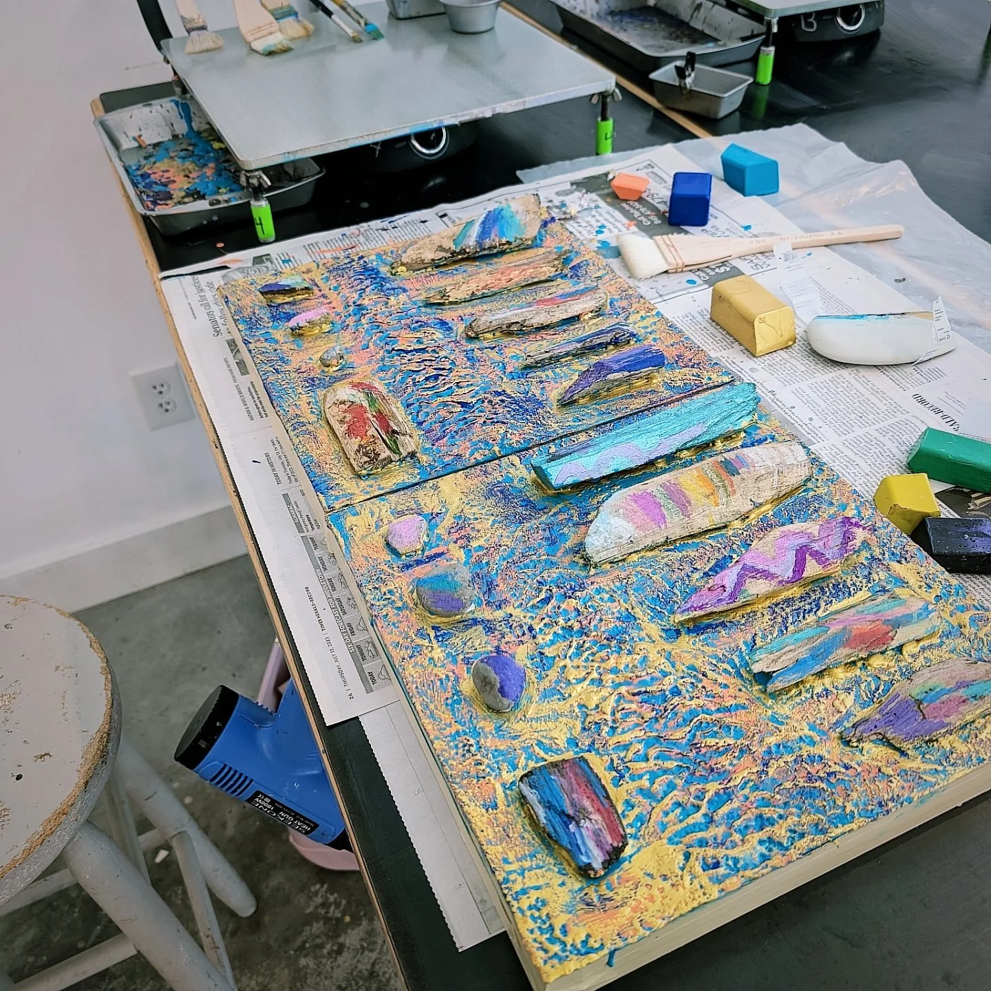 A mixed media art piece featuring colorful, textured stones on a canvas covered with yellow and blue paint. Art supplies like paintbrushes, sponges, and pastel sticks are scattered around. The workspace is covered with newspaper.