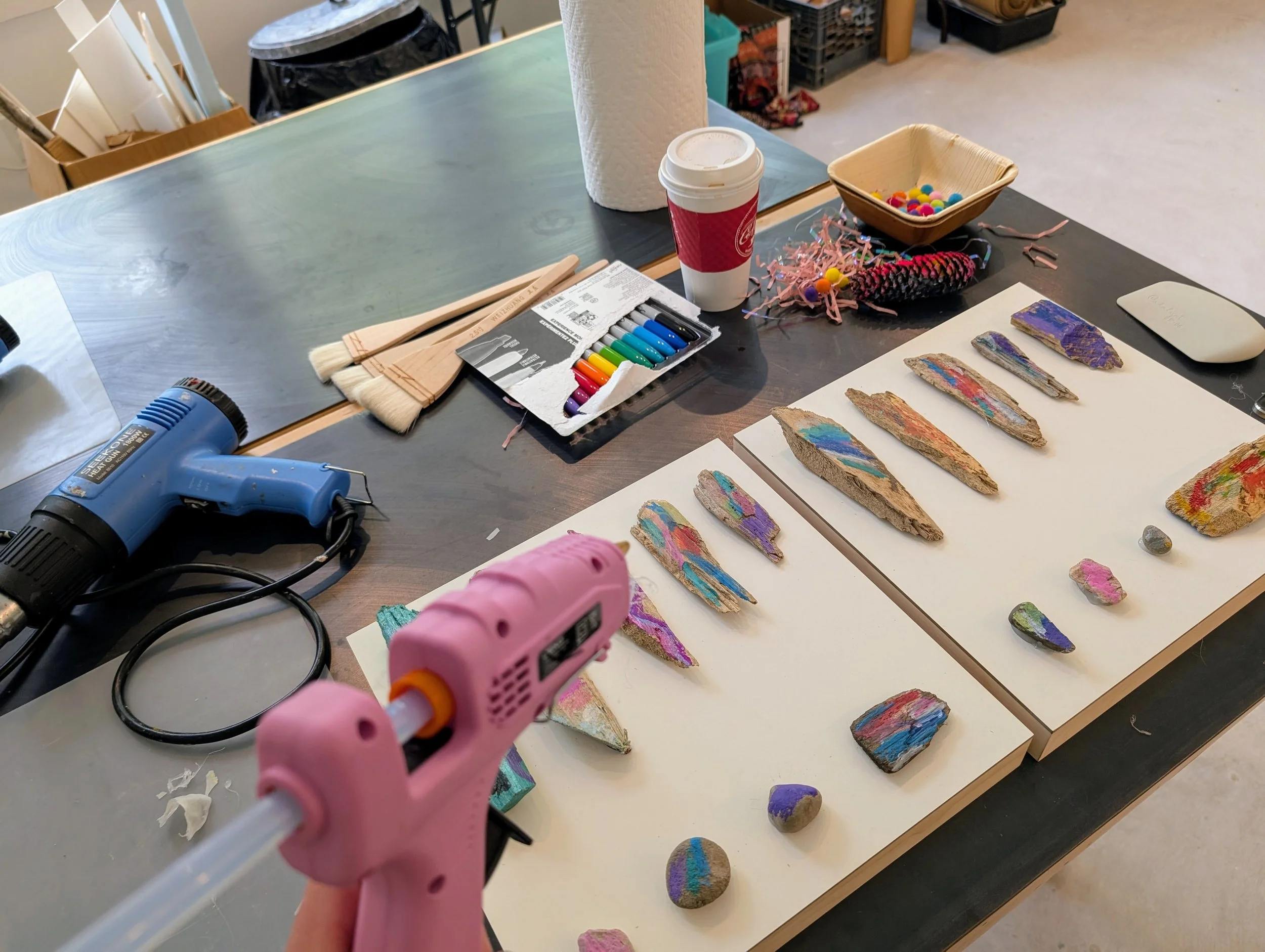 A workspace with art supplies, a hot glue gun, glue sticks, paint brushes, a set of colored markers, and painted rocks arranged on two white boards. There is a paper cup with a lid, a basket of small colorful pom-poms, and a piece of pink shredded pa