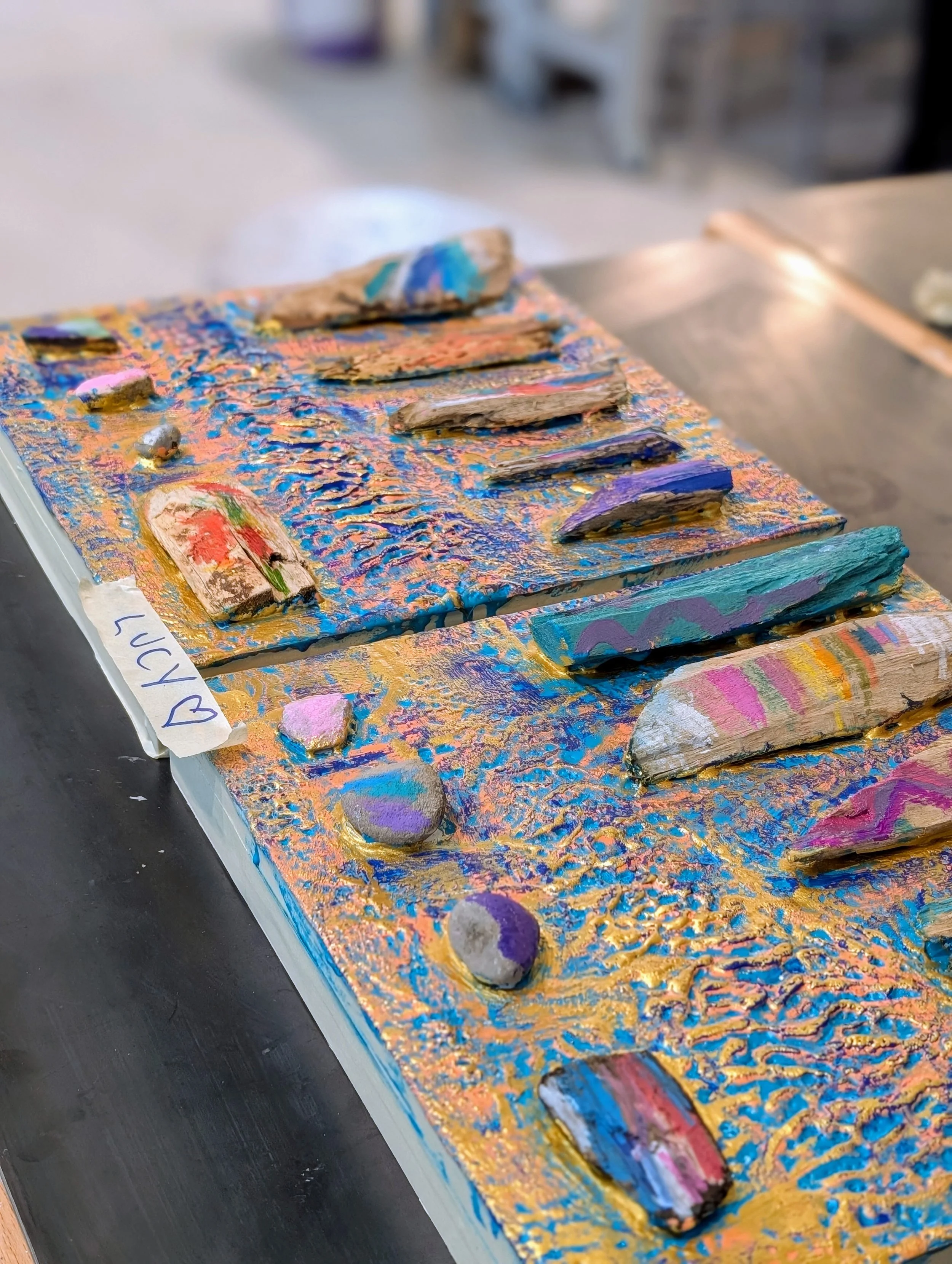 Colorful textured art piece with various painted rocks and wooden pieces arranged on a canvas, with a handwritten note saying 'LUCY' with a heart symbol.