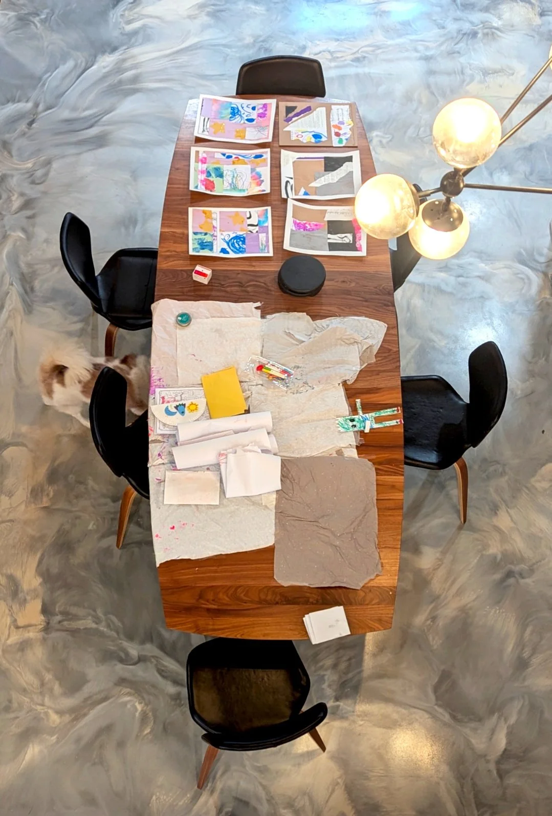 Overhead view of a wooden conference table with art projects, paper, and art supplies scattered on the surface. Four black chairs surround the table, and a fluffy cat is near the bottom left corner. The background shows a gray marble floor and a mode