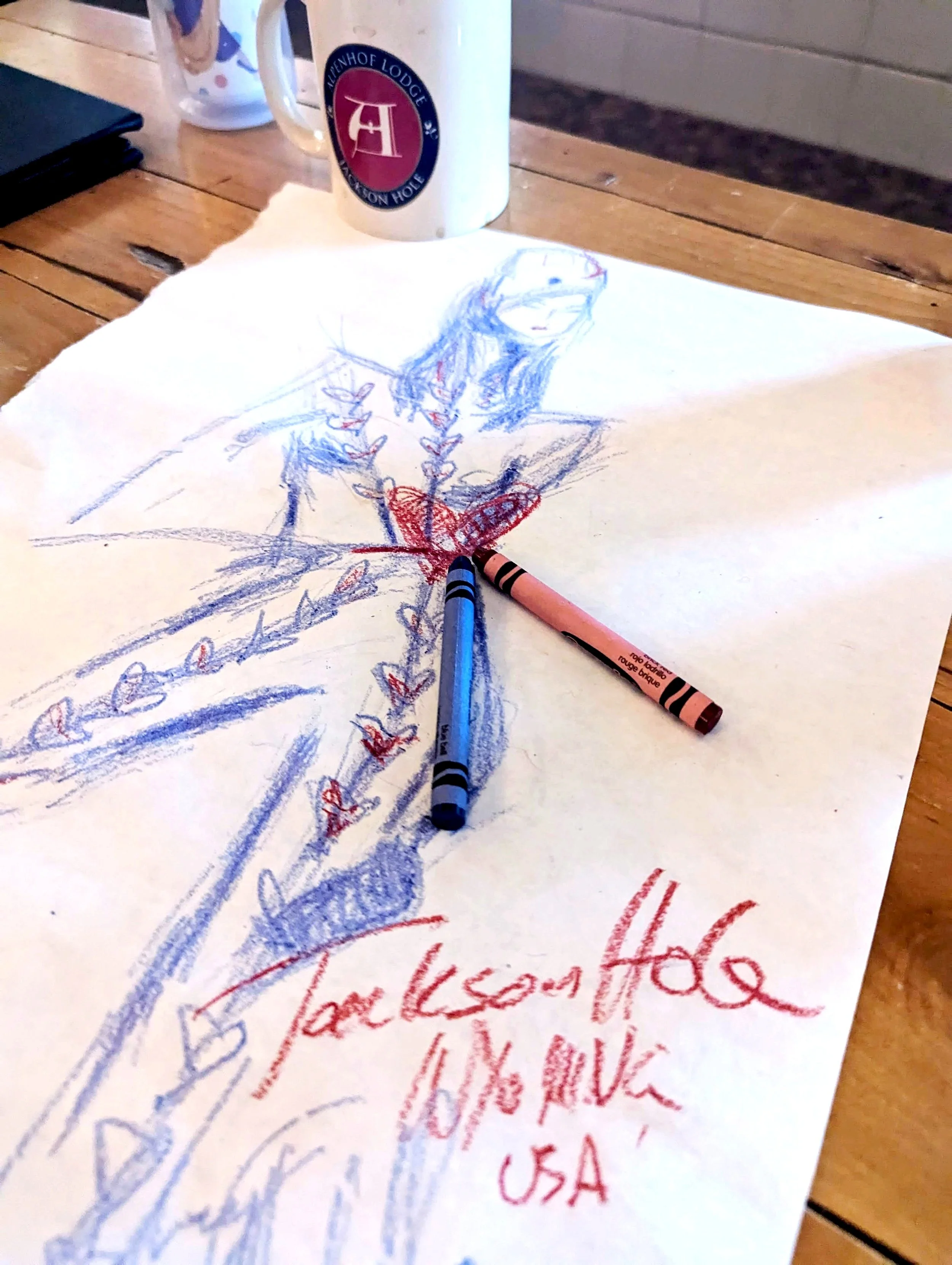 A drawing of a woman, partially sketched, on a sheet of white paper. Two crayons, one red and one blue, are placed on the paper. In the background, there is a white mug with a logo and a ceramic container, all on a wooden table.