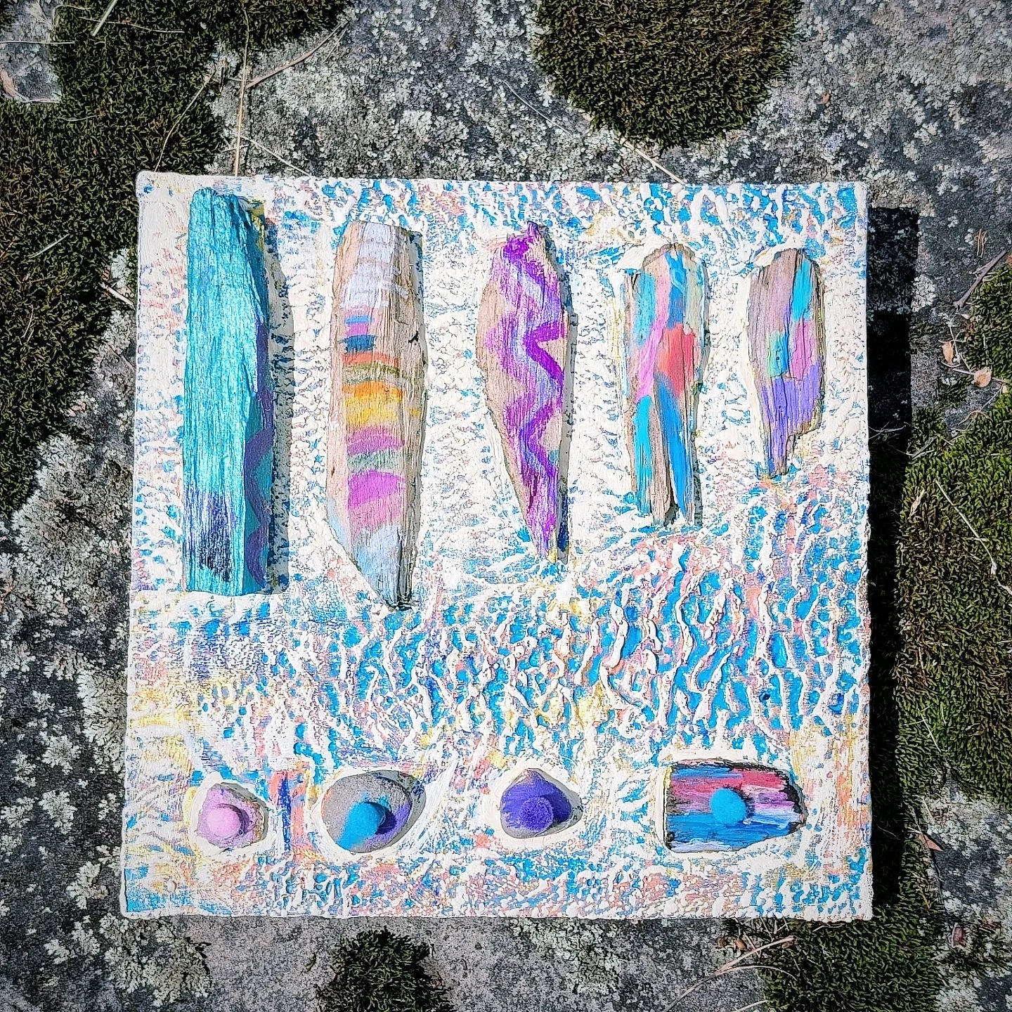 Colorful abstract painting on a white textured canvas with five vertical, textured brushstrokes above four circular shapes, outdoors on a rocky surface surrounded by small green plants.