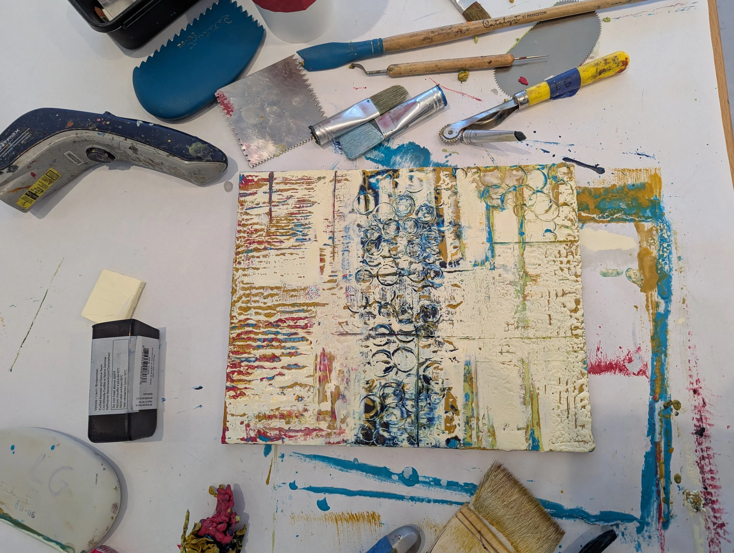 Abstract textured artwork with circular patterns on a canvas, surrounded by art supplies like paintbrushes, razors, spray paint cans, a box cutter, and colorful paints on a white table.