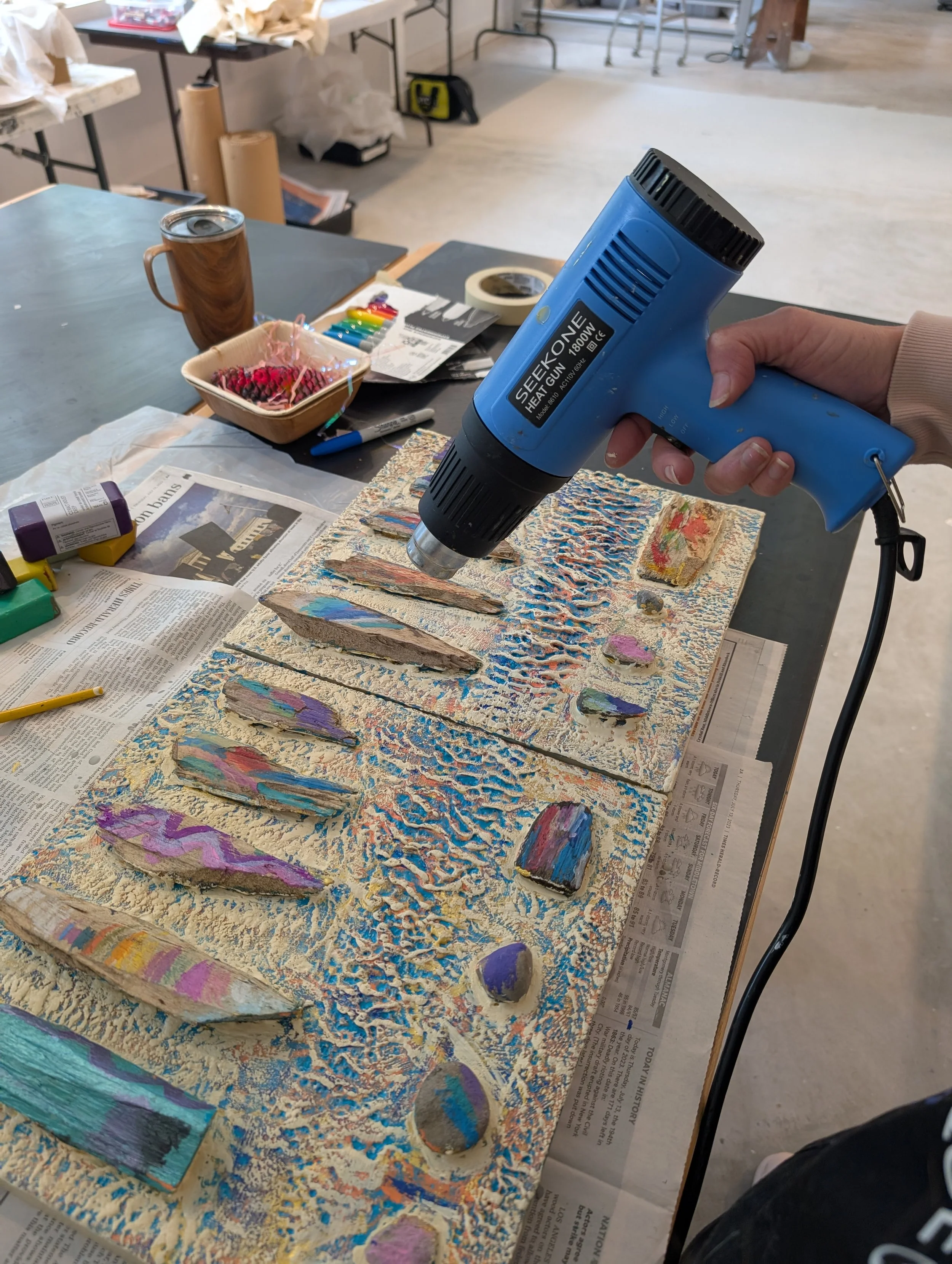 Person using a heat gun to craft or paint on a textured artwork involving stones on a table covered with newspaper, with art supplies and tools visible in the background.