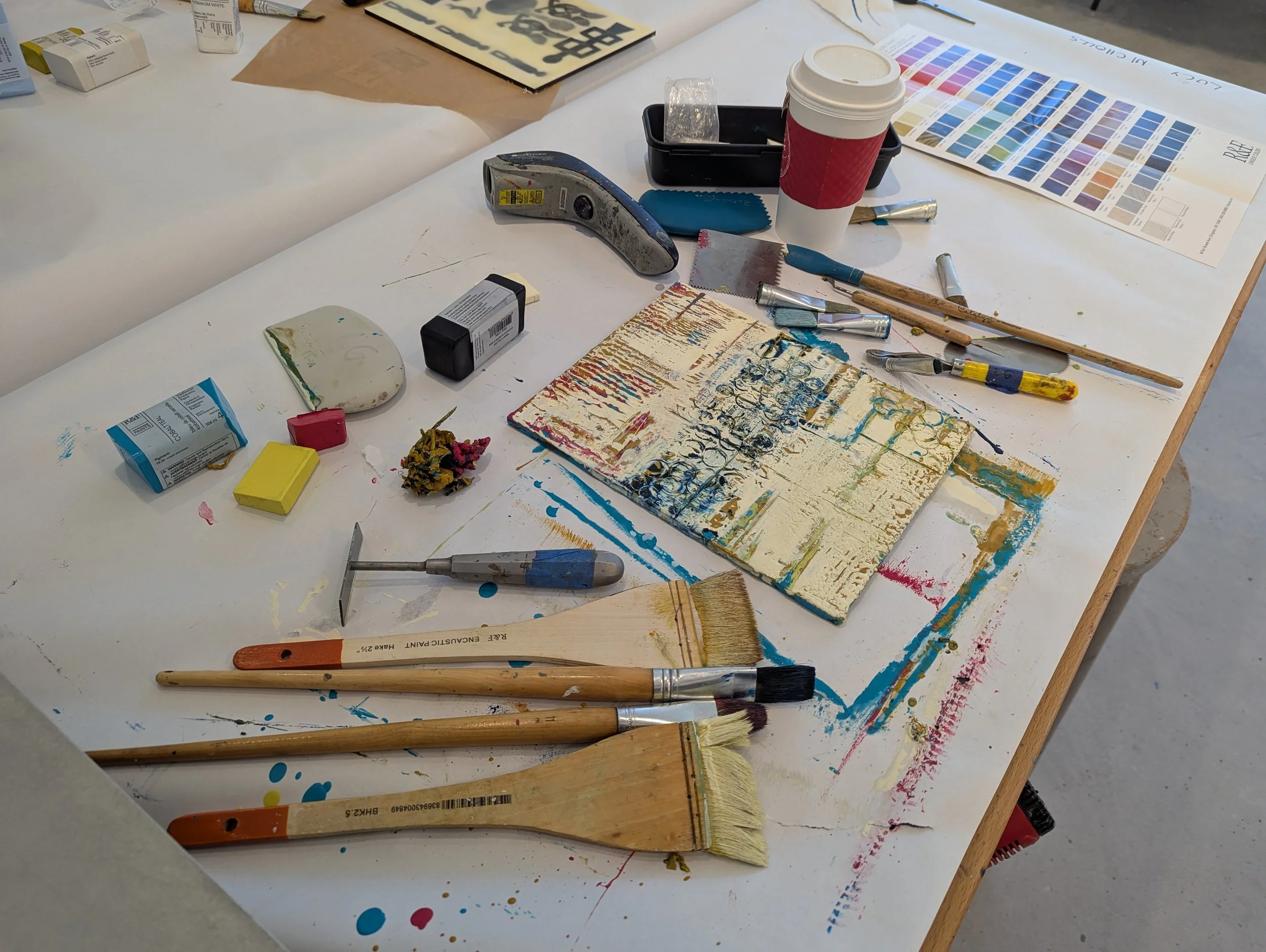 Art workspace with paintbrushes, paint, palette knife, and a cup, surrounded by paint swatches, a textured canvas, and various art supplies.