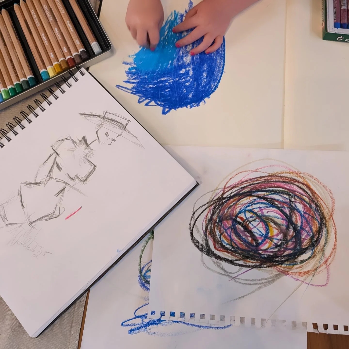 Child's hands drawing with crayons on paper, creating a blue circular scribble. Nearby are colorful crayon sets, a sketchbook with rough animal sketches, and abstract scribbles on separate sheets of paper.