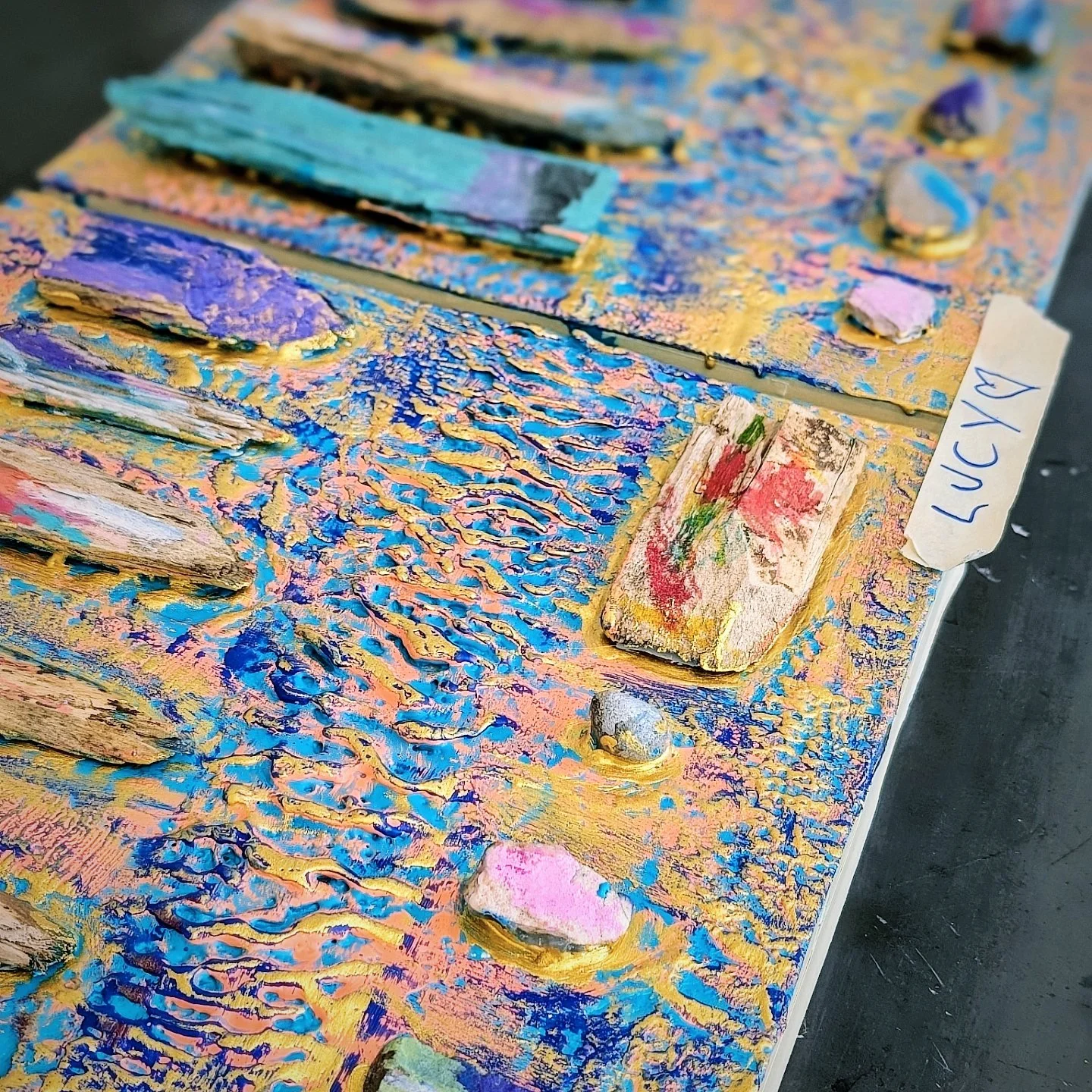 Close-up of textured, colorful abstract artwork with thick paint strokes in purple, pink, blue, green, and gold, labeled with a sticky note that says 'LUCY' and a heart symbol.