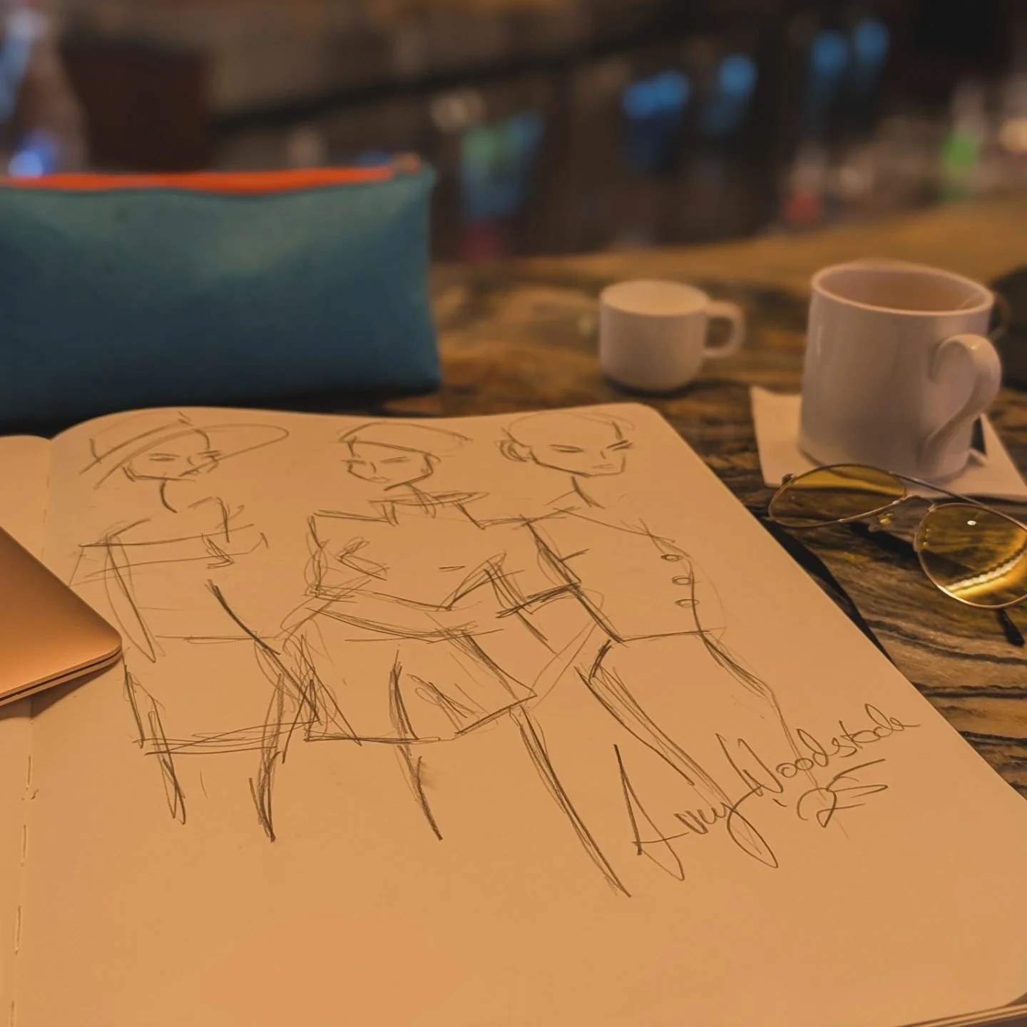 Sketch of four women in stylish outfits on a notebook page, placed on a table in a cozy cafe setting with cups, sunglasses, and a blue bag nearby.