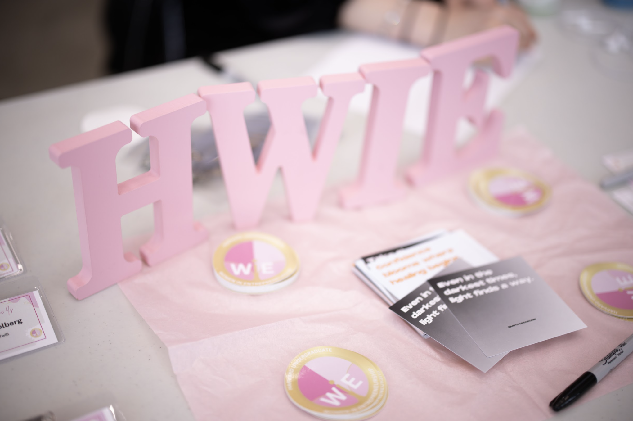 Pink decorative letters spelling 'WE' on a table with pink and gold circular stickers and pamphlets at a table.