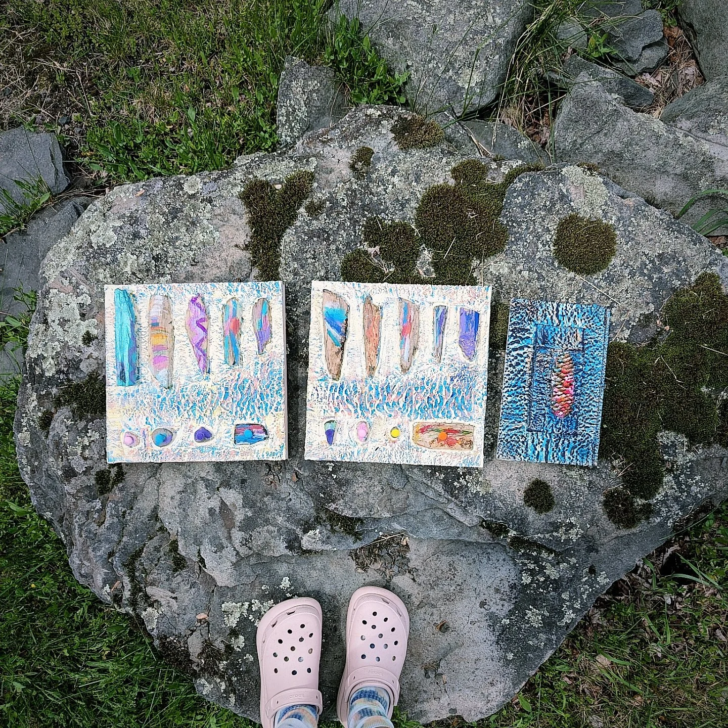 Three colorful watercolor paintings placed on a large mossy rock outdoors, with a pair of pink Crocs at the bottom and green grass around.
