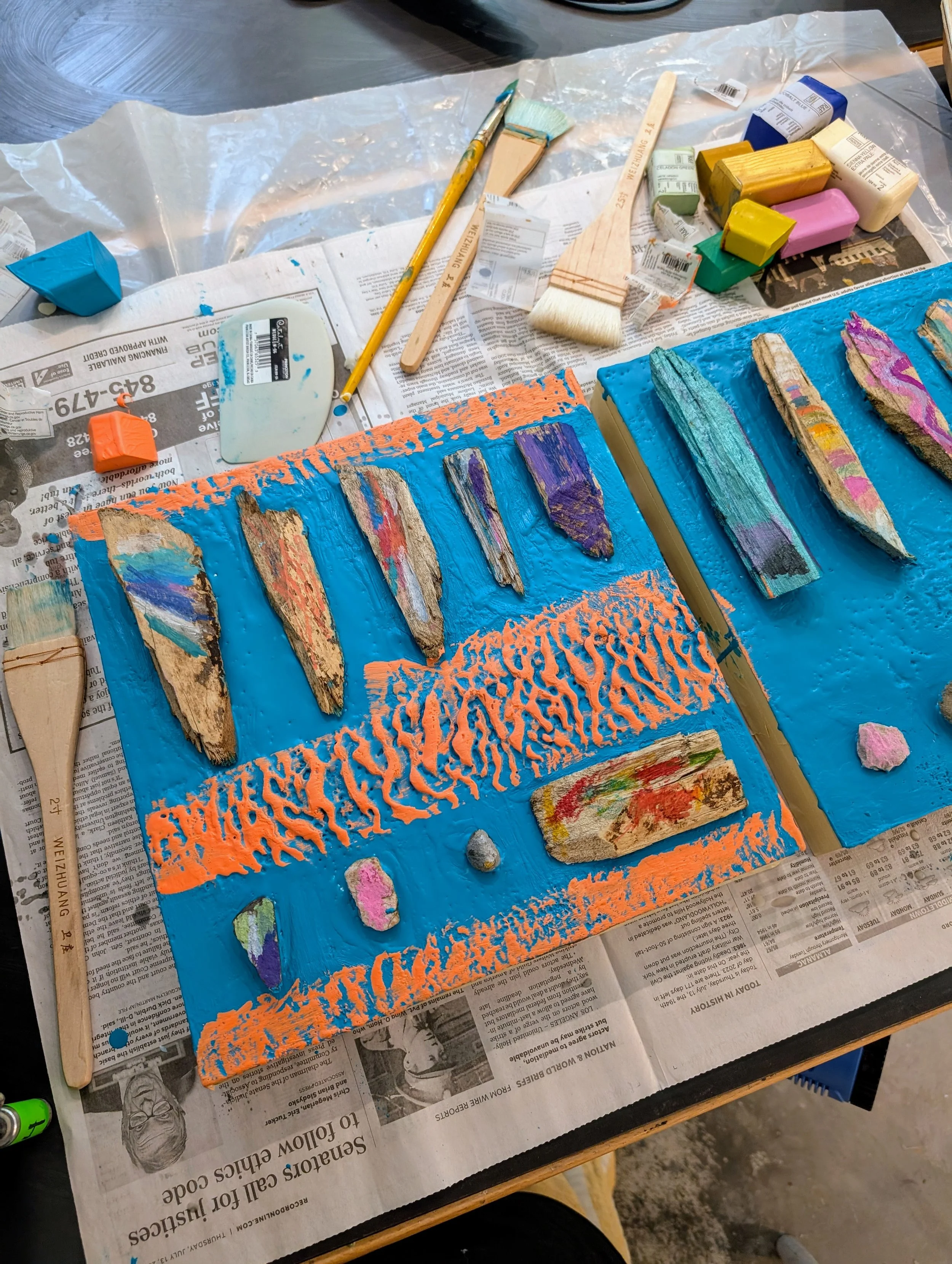 A workspace with painted wooden pieces on a blue painted surface, with various painting tools and art supplies around, including brushes, paint, and sponges.