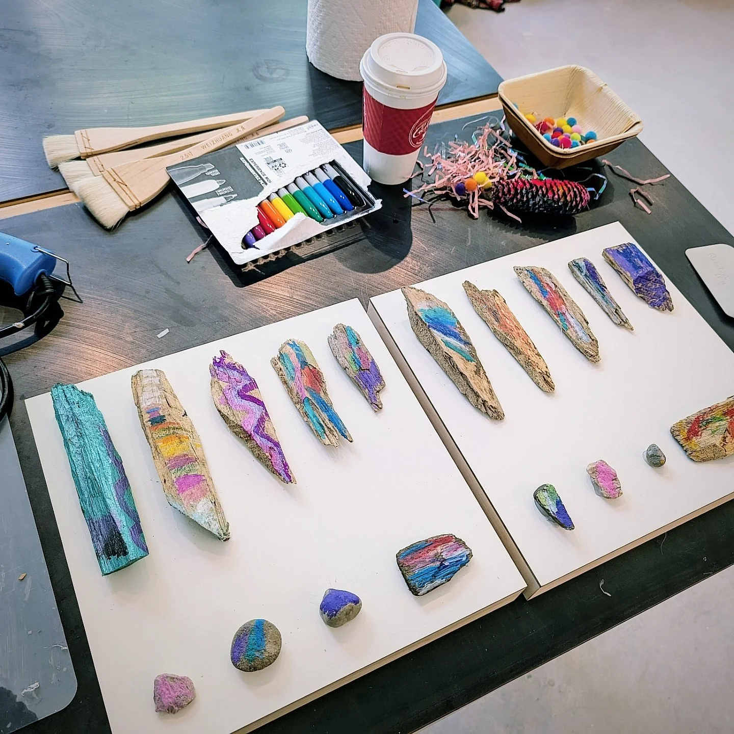 Colorful painted rocks arranged on white boards, with art supplies, such as paintbrushes, markers, and decorative items, on a table.
