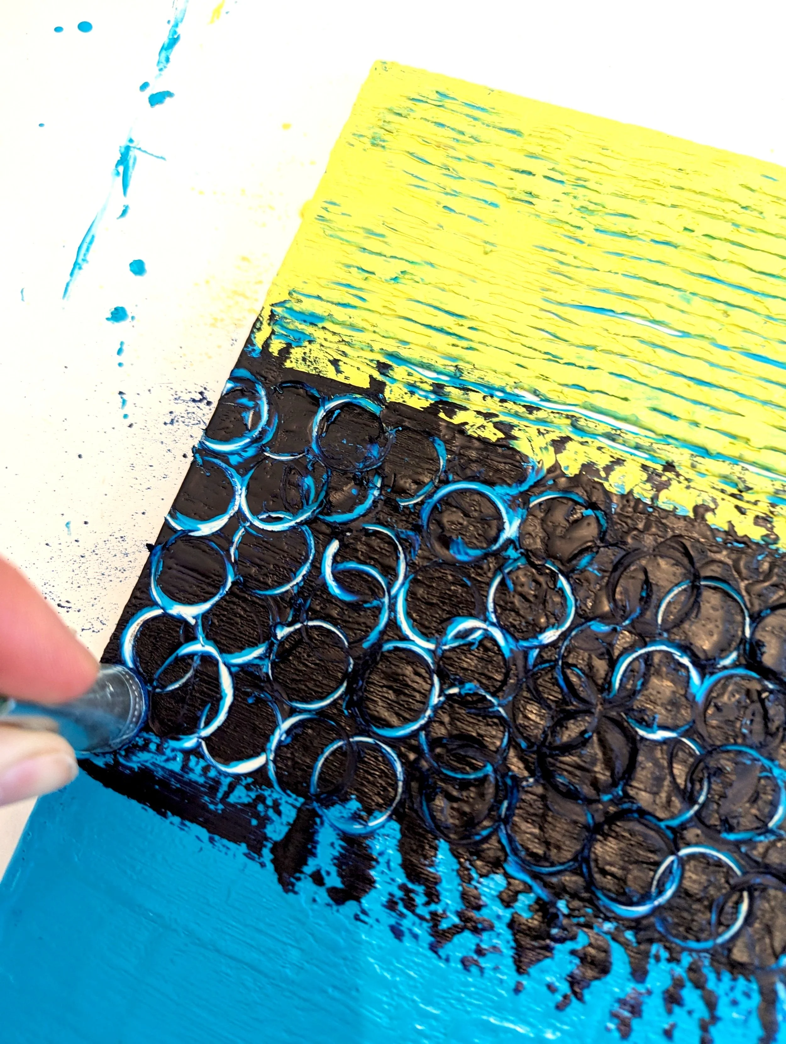 Close-up of a hand spray painting a piece of textured artwork with black, yellow, and blue colors. The artwork features a section with small circular patterns and abstract textures.