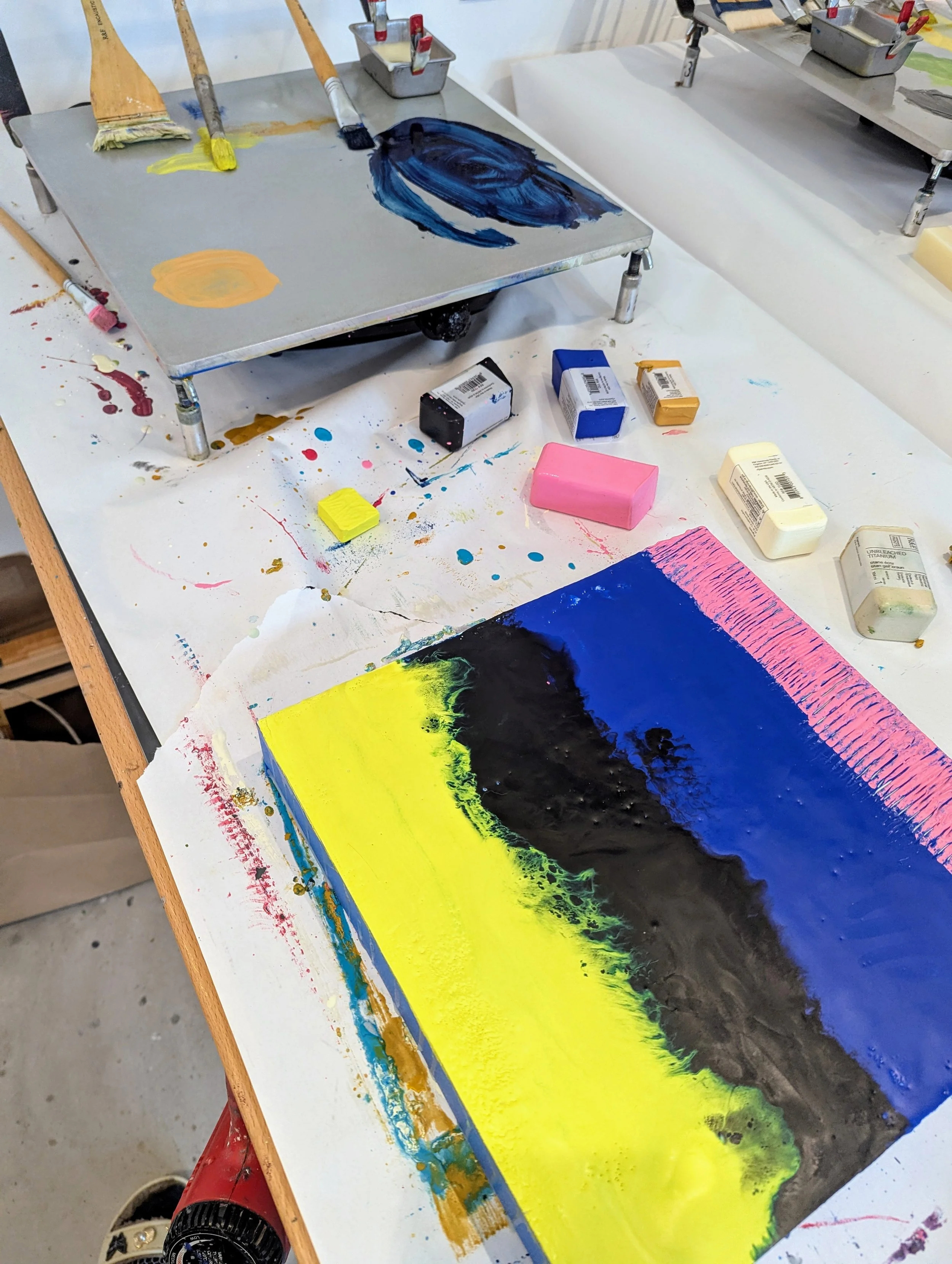An art workspace with a canvas featuring a colorful abstract landscape painting with yellow, black, blue, and pink hues. Various paint bottles, brushes, and tools are scattered around, with some paint splatters and drips on the table.