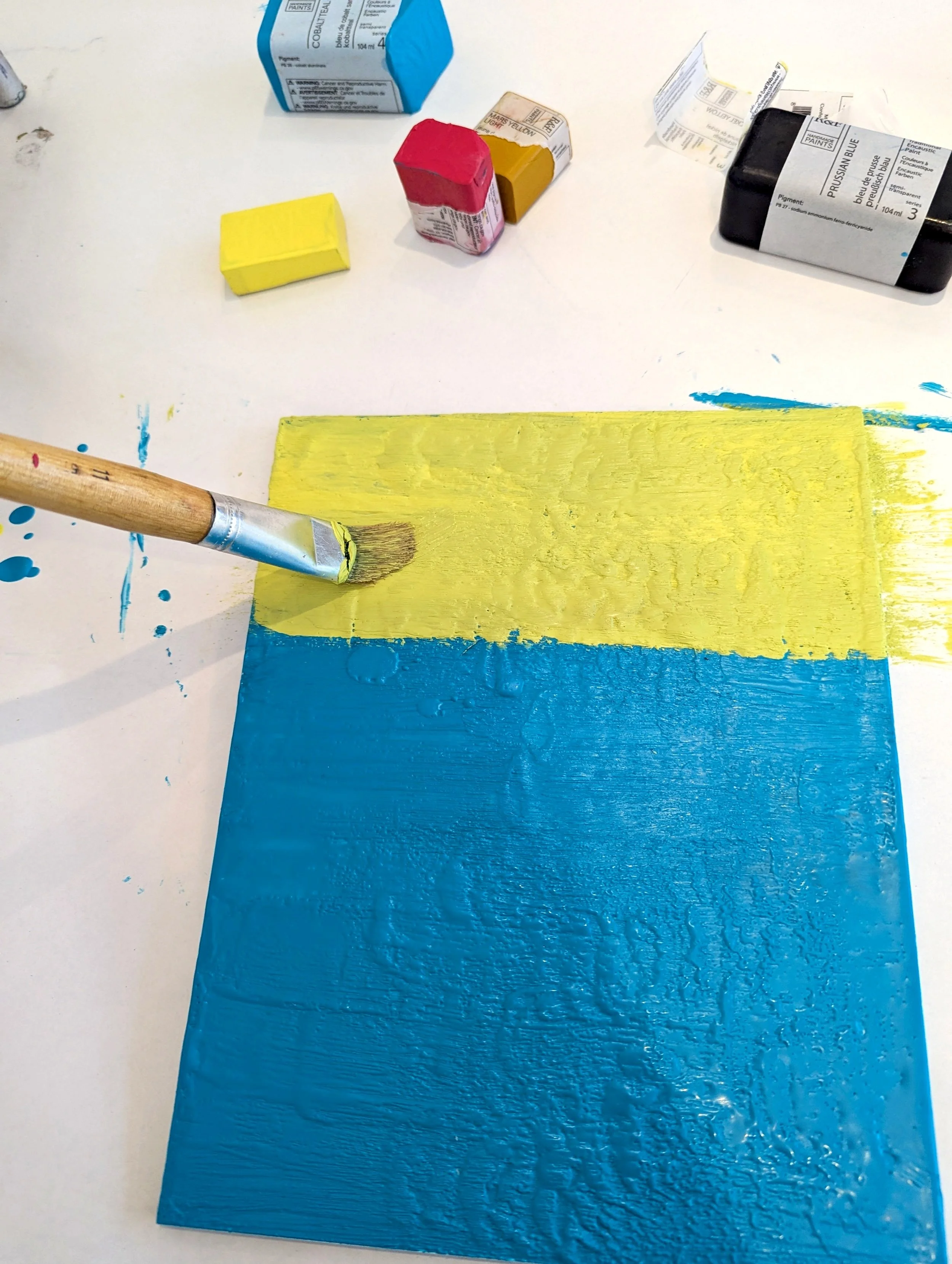 A person is painting a wooden board with yellow and blue paints, using a paintbrush. The table has paint supplies, including small paint containers, scattered around.