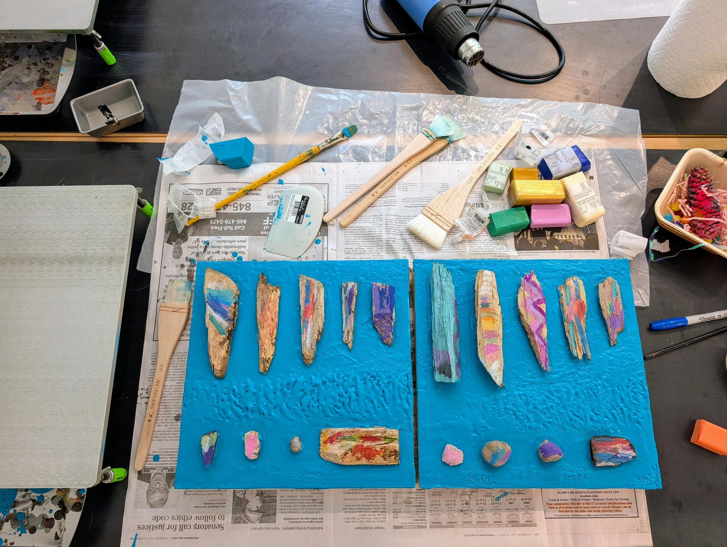 Colorful painted rocks and stones on blue textured foam boards, surrounded by art supplies like paint, brushes, and sponges, on a work table covered with newspapers.