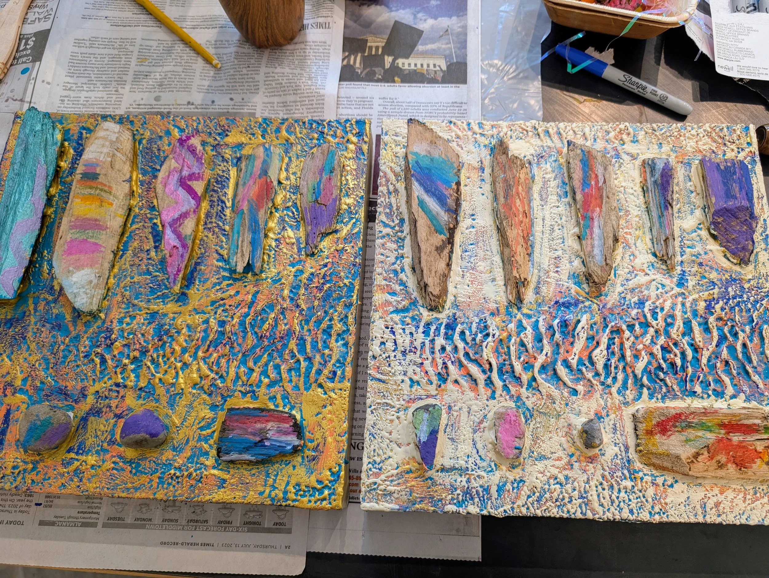 Two textured art panels with painted rocks and wooden pieces on a table covered with newspapers and art supplies.