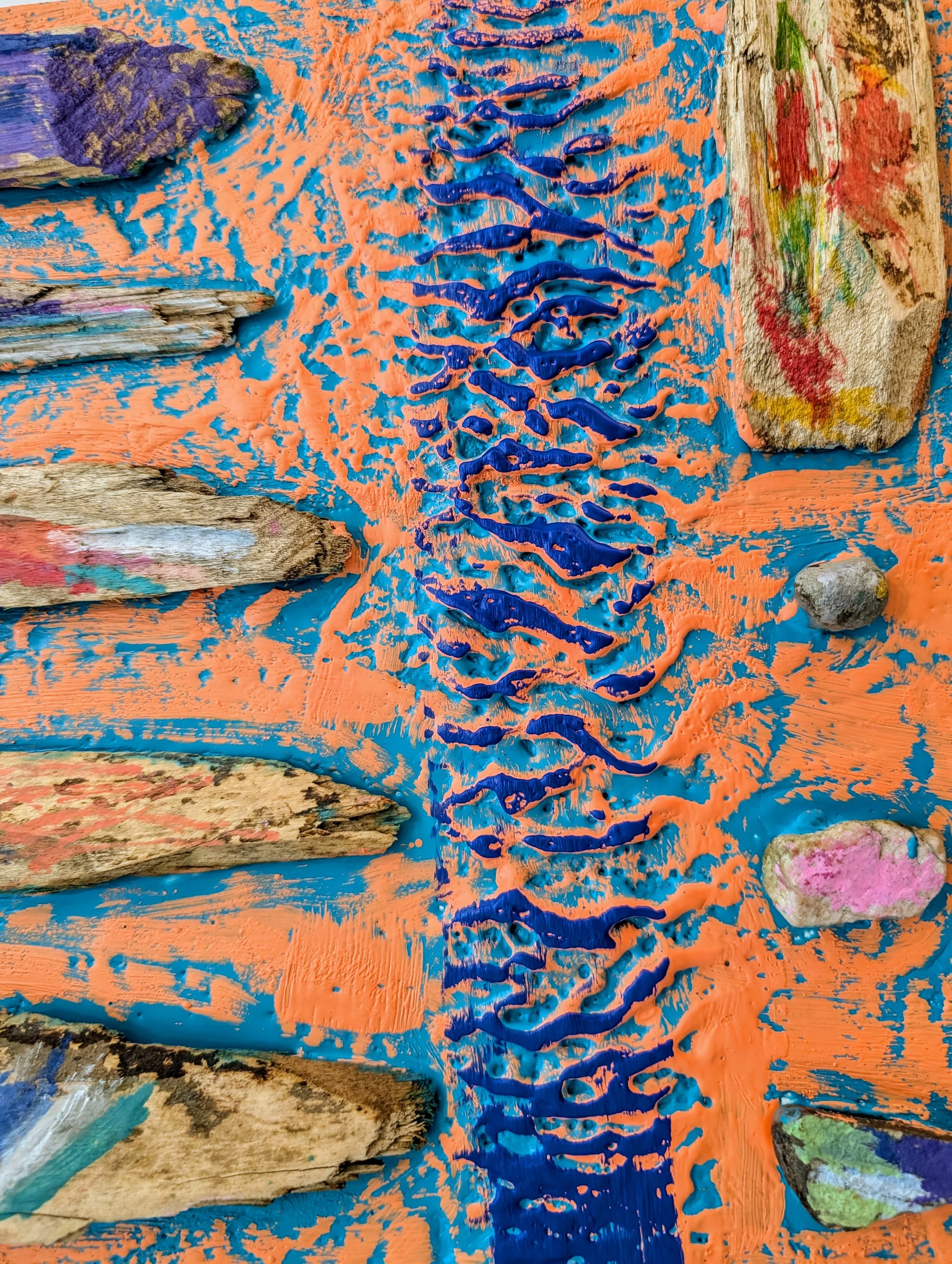 Close-up of an abstract textured artwork with painted rocks and pieces of wood on a vibrant orange and blue background.