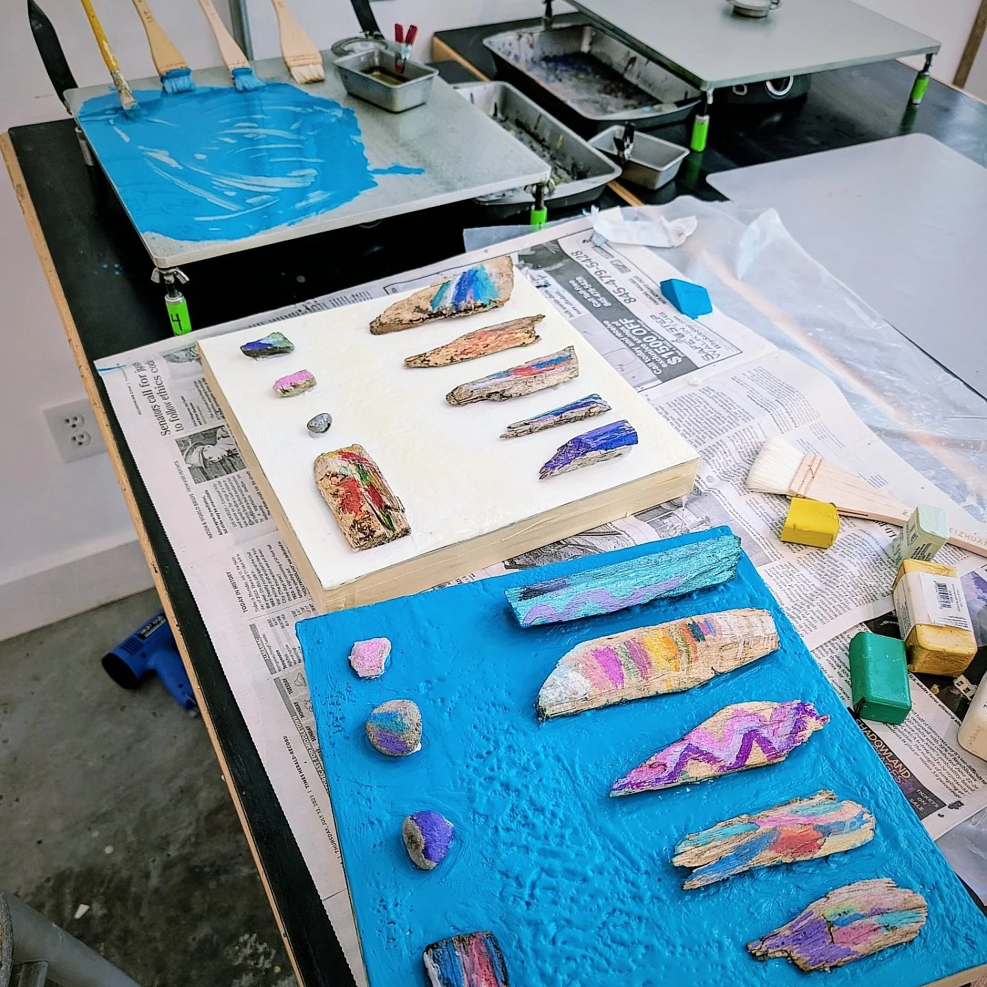 An art studio workspace with painted rocks and wood pieces on two boards covered in bright blue and cream paint. Art supplies and newspapers are scattered around.