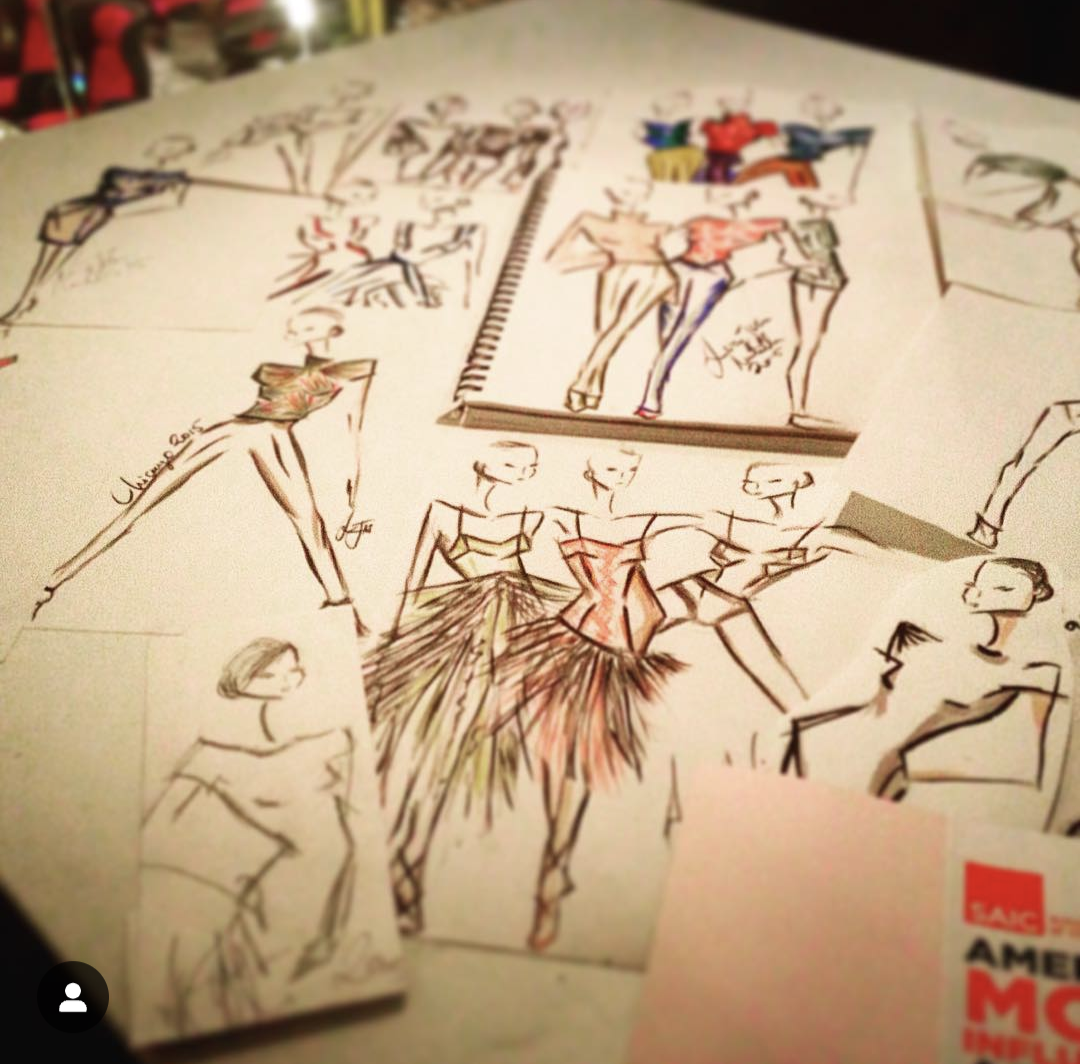 Sketches of women in fashionable outfits, some with colorful details, laid out on a table with art supplies in the background.