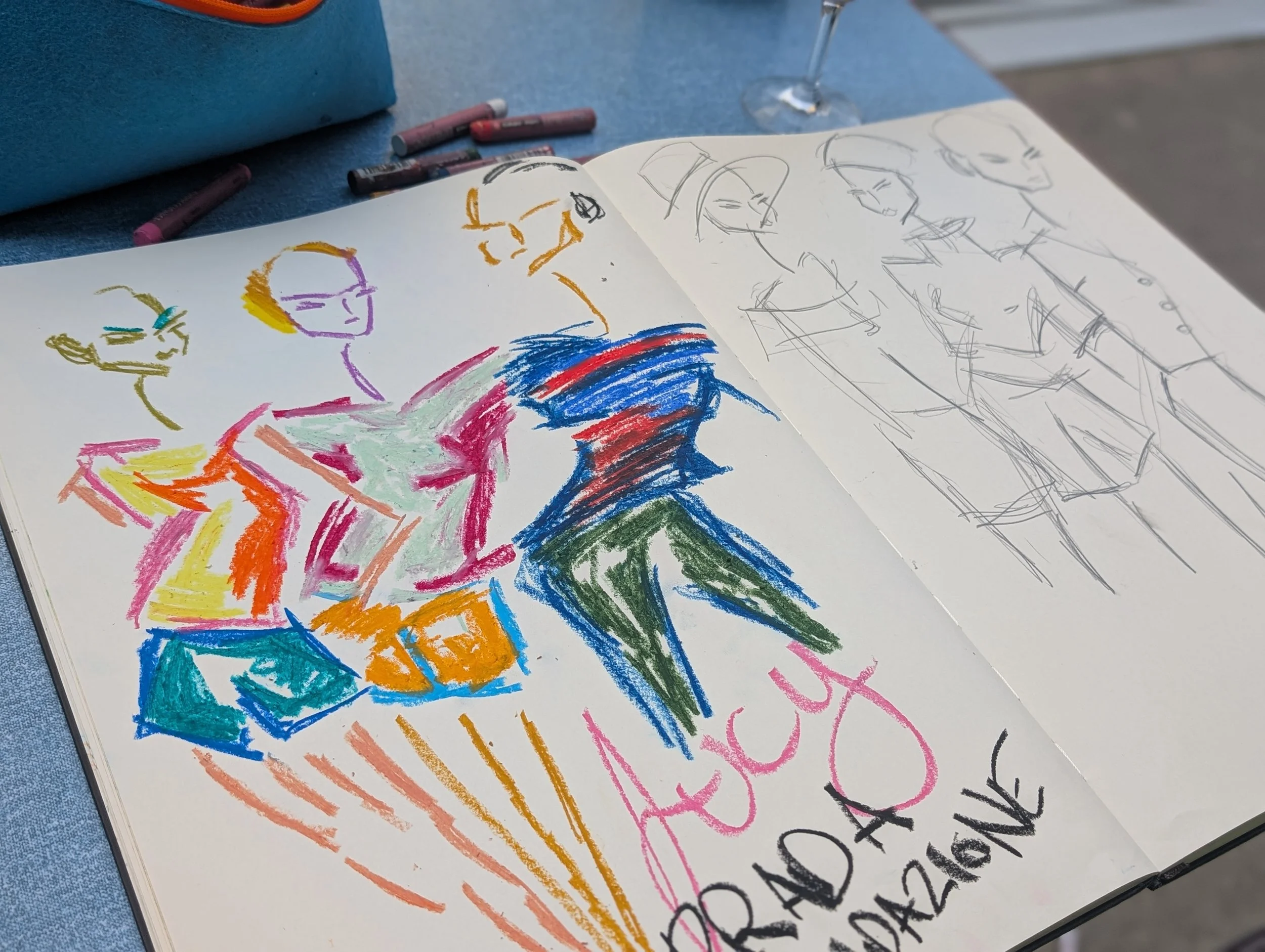 Sketchbook with colorful fashion illustrations on the left page and black and white sketches on the right page, placed on a blue table with colored pencils nearby.