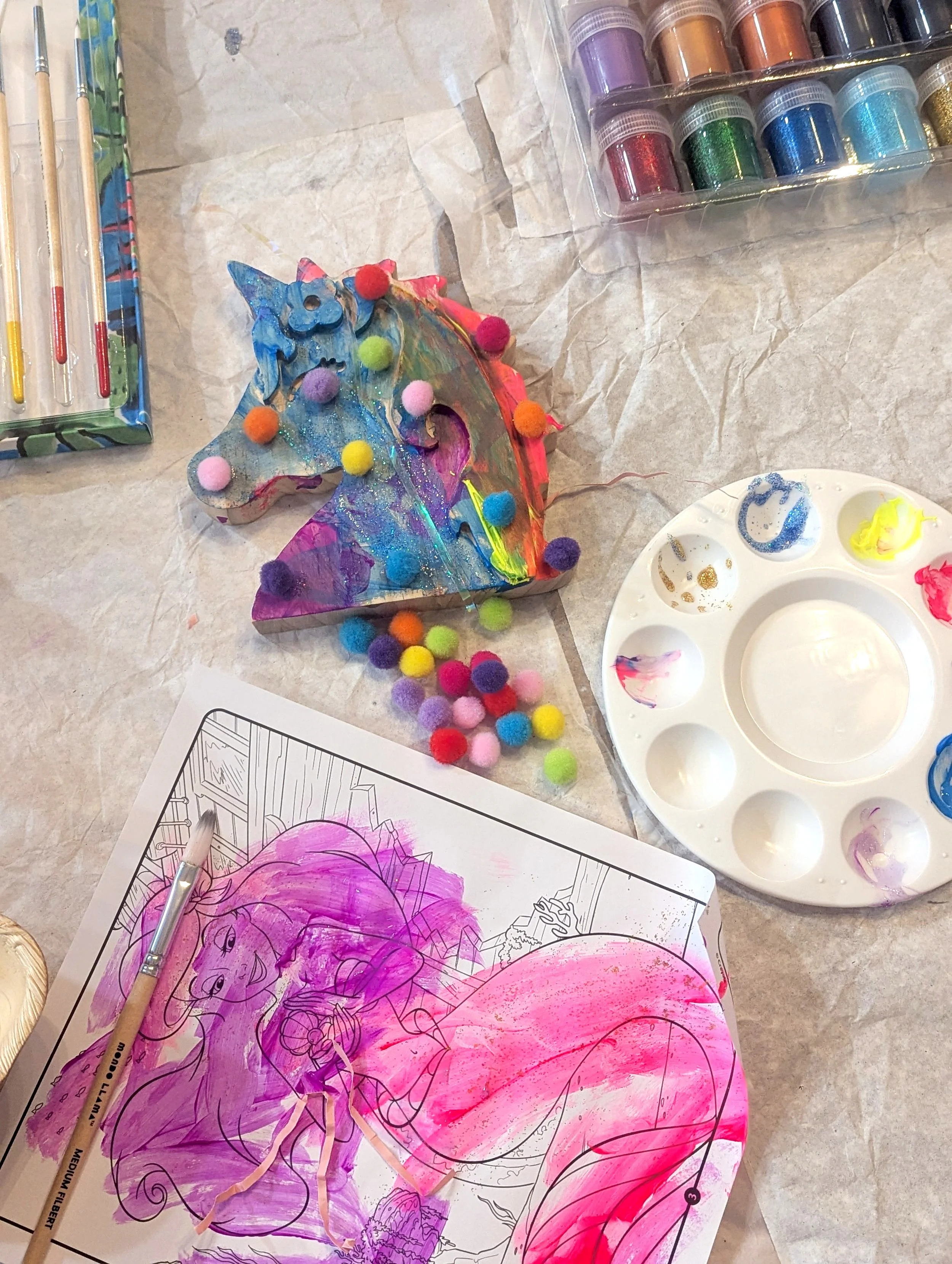A colorful unicorn craft with pom-poms, watercolor paints, a paint palette, and a coloring page on a table.