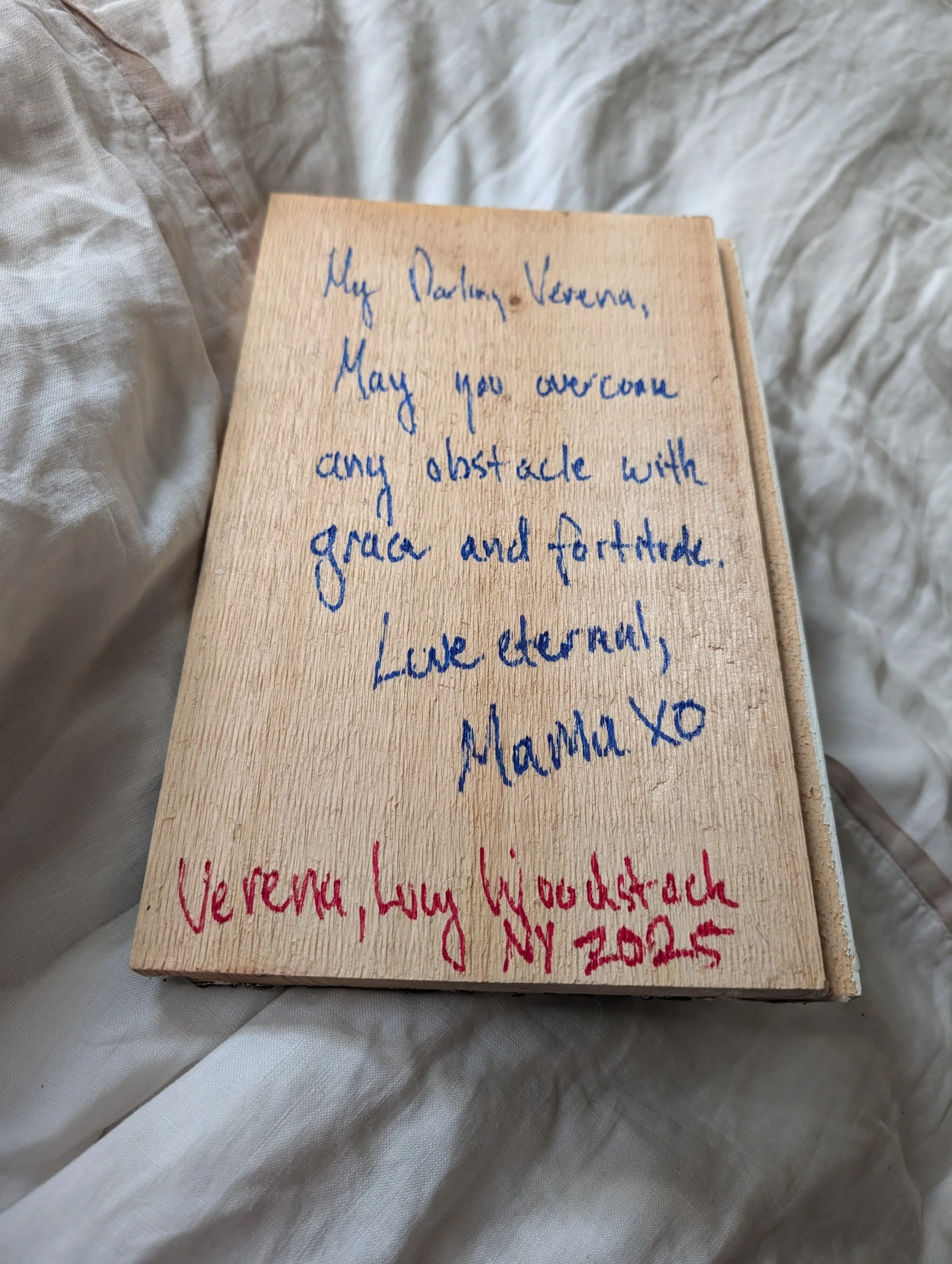 A wooden plaque with a handwritten note in blue and red ink. The note contains a message to Vera and Lucy from Mama, expressing love, hope for strength and grace, and includes an address in New York, 2025.