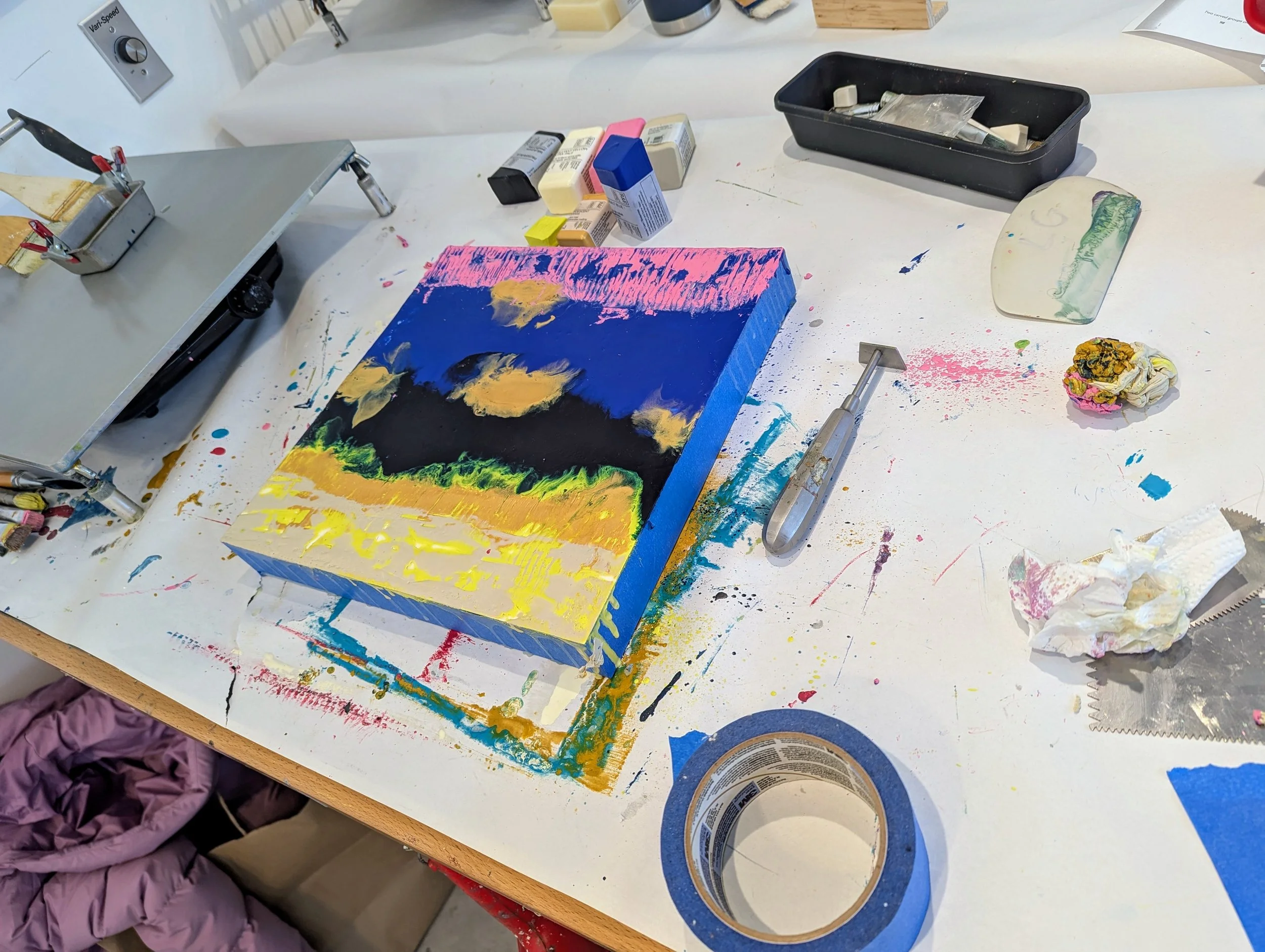 A colorful landscape painting of a sunset or sunrise on a small canvas, surrounded by art supplies including tape, crumpled tissue, a paint spatula, and paint bottles on a messy white worktable.
