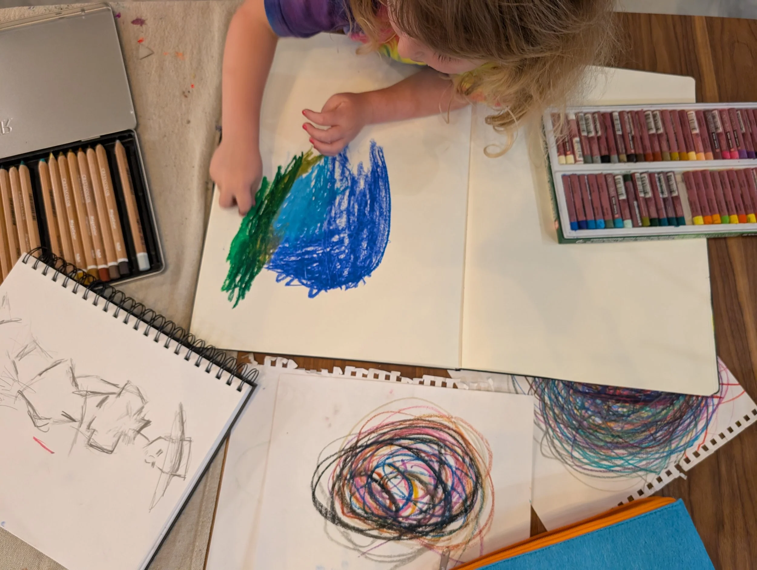 Child drawing with crayons on a large sheet of paper, with other colorful scribbles and sketches around, on a wooden table.