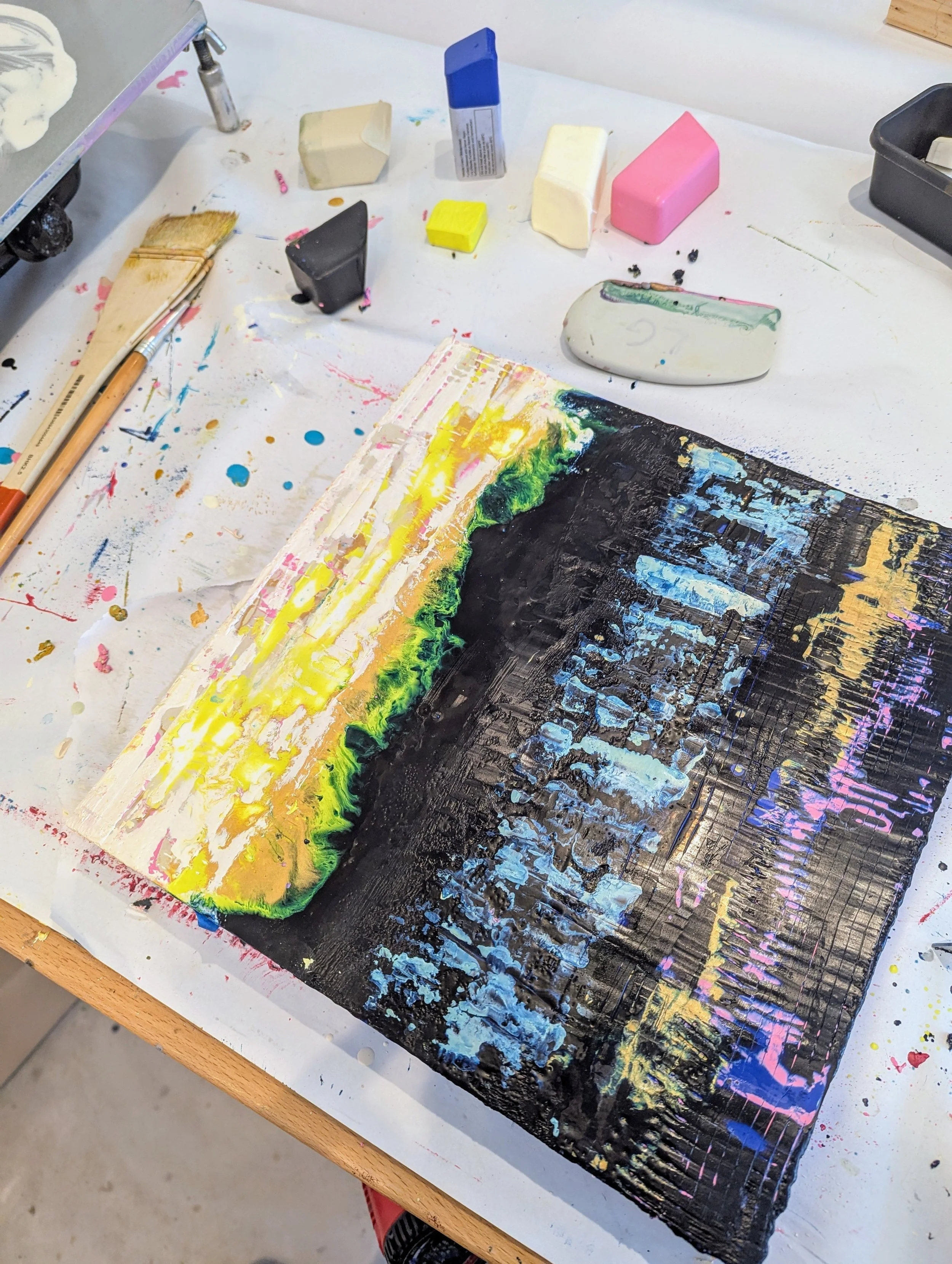 Abstract mixed media artwork on a canvas with bright yellow, green, black, and blue colors, surrounded by art supplies including erasers, paintbrushes, and pastel blocks.