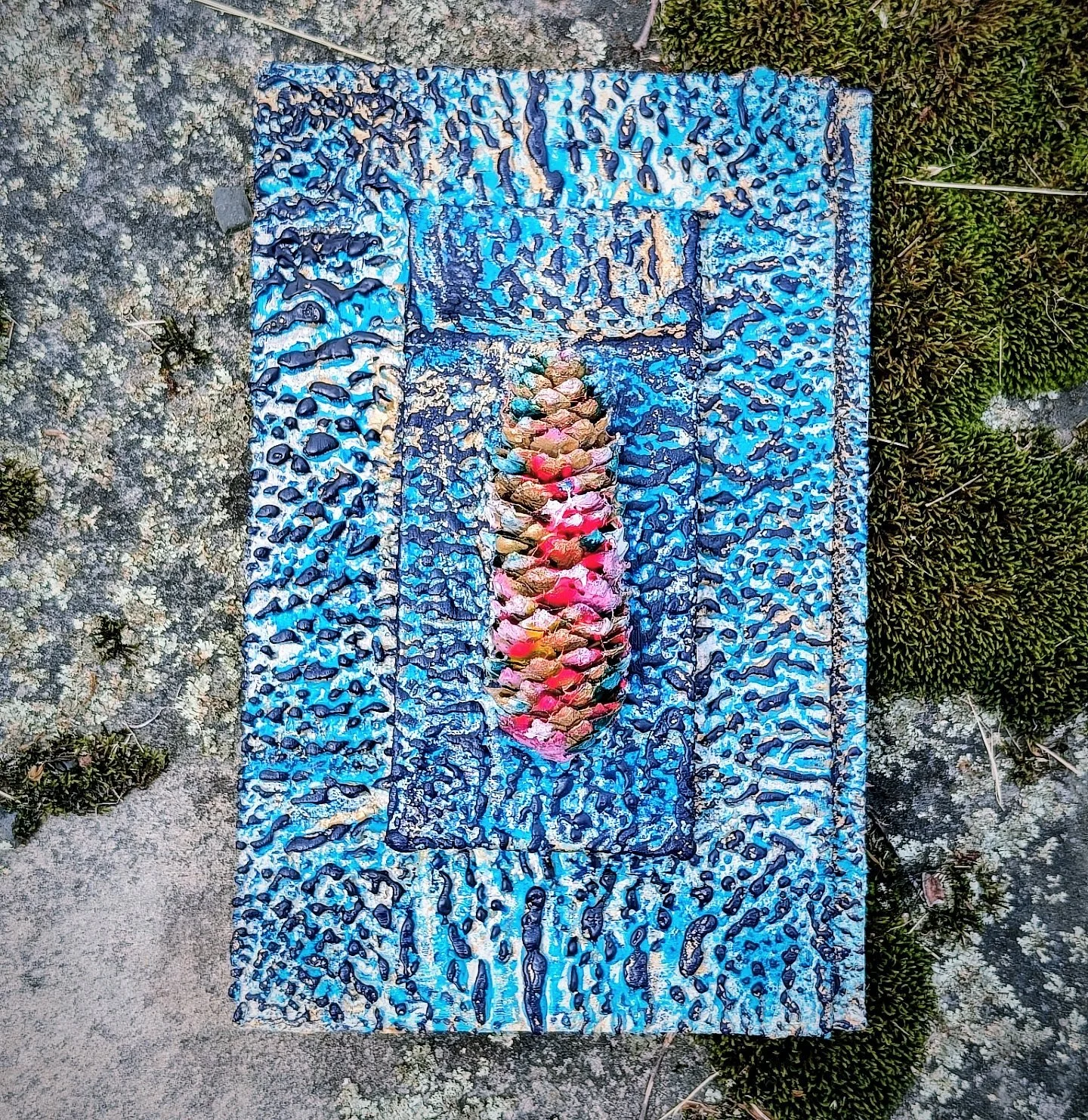 Colorful abstract textured artwork on a rectangular canvas, featuring a central cone shape with multi-colored, layered scales, surrounded by a vivid blue and black patterned background, placed on a stone surface with some greenery nearby.