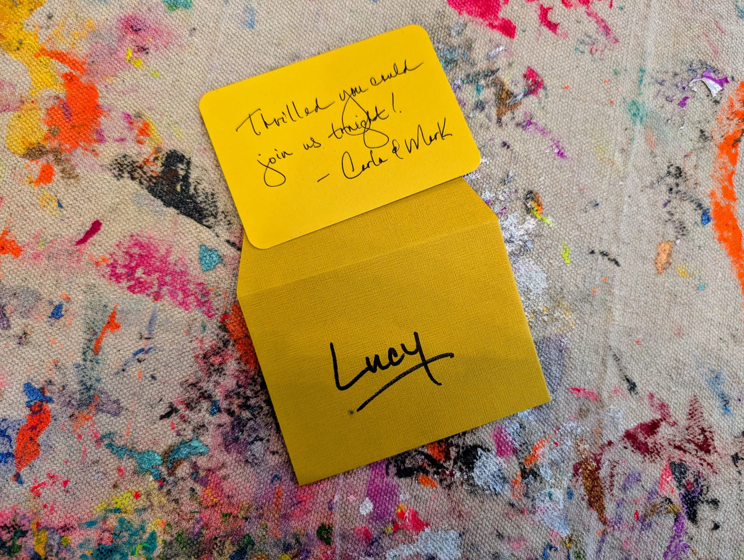 A yellow notepad with a handwritten note and signature on a colorful, paint-splattered surface. The note reads: 'Thrilled you could join us tonight! - Cara & Mark' and the signature is 'Lucy'.