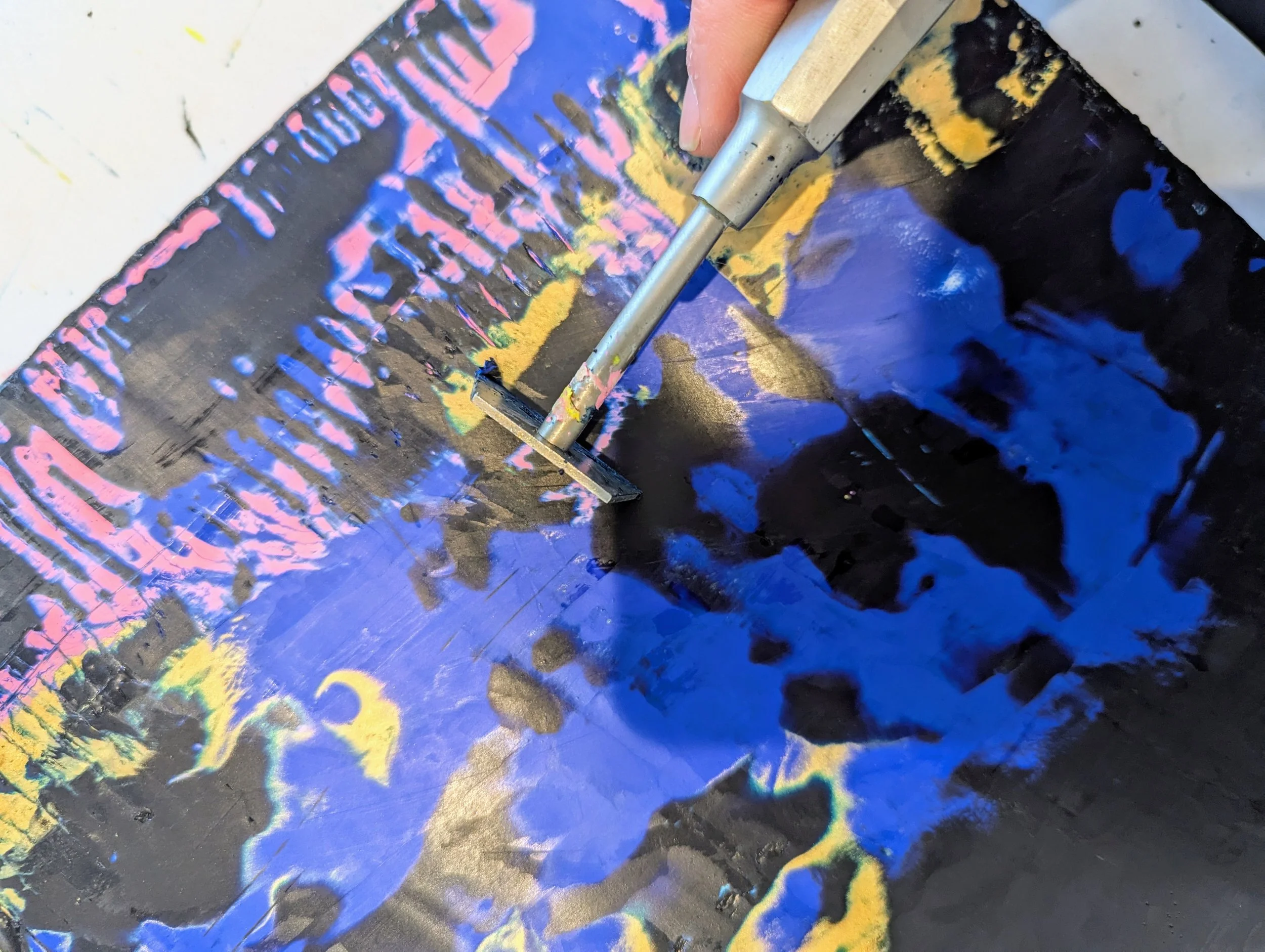 A close-up view of a glass painting with acrylic paints in pink, blue, yellow, and black. A hand is using a metal painting tool to spread or scrape paint on the glass surface.