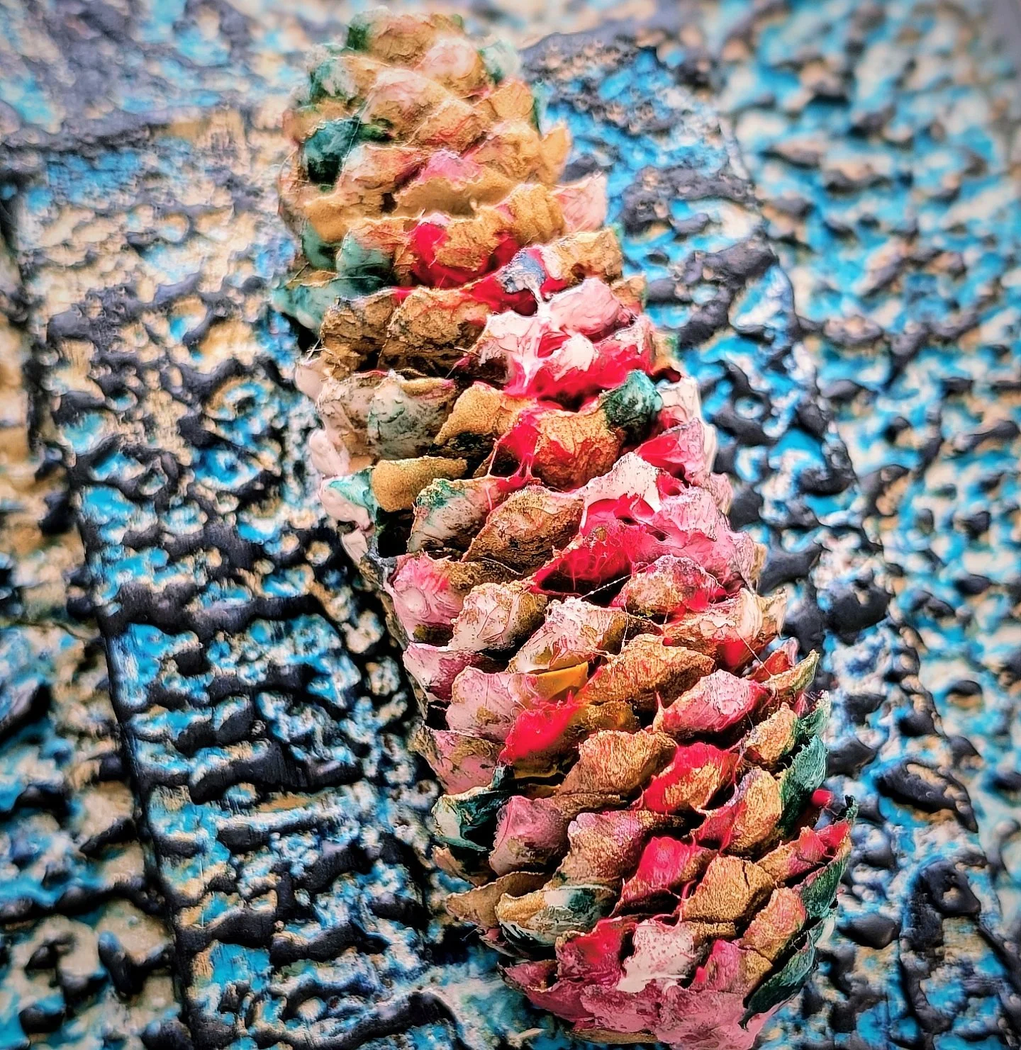 Close-up of a pine cone with red, pink, green, and brown colors, set against a textured blue and black background.