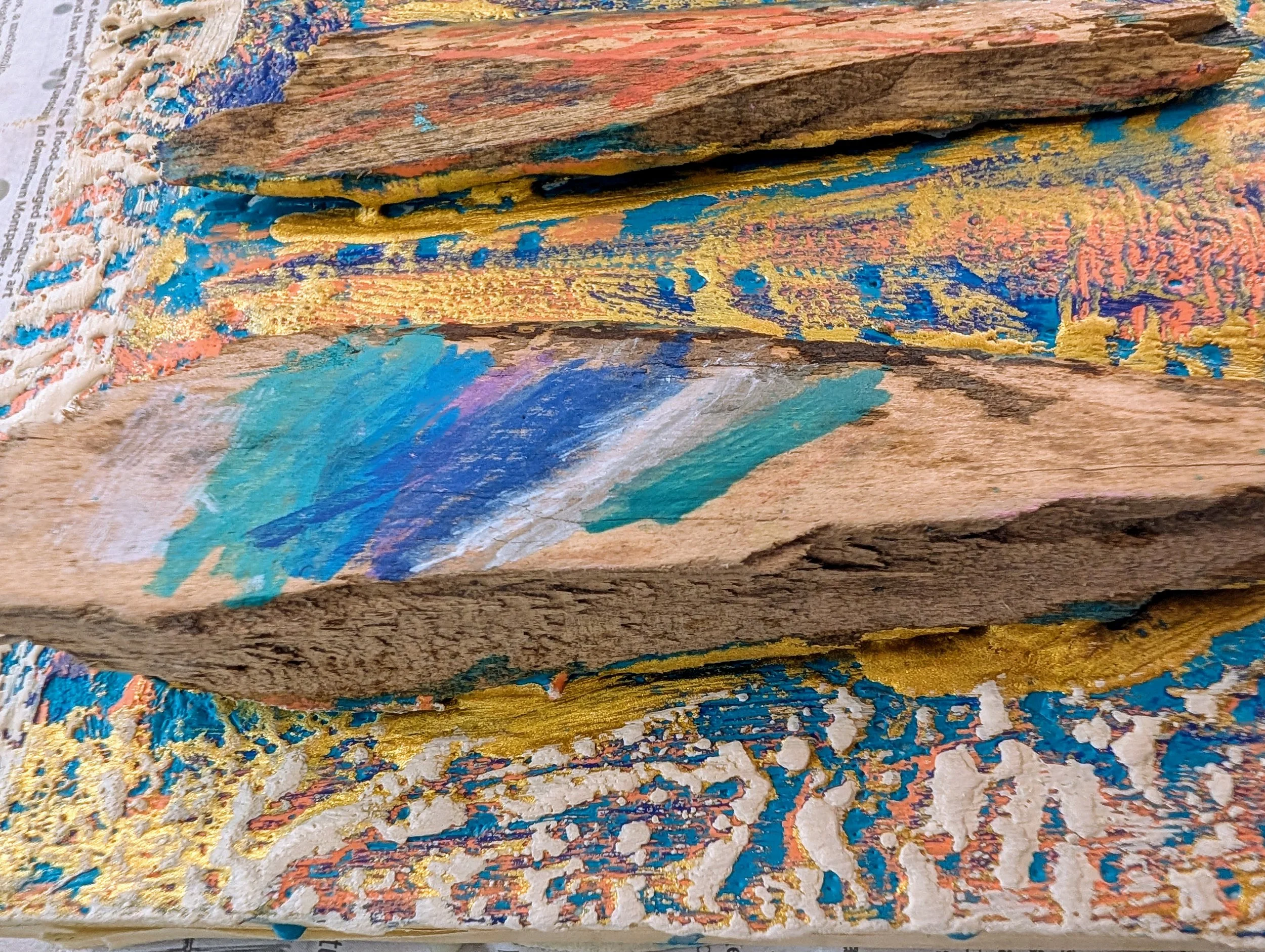 Close-up of a painted wooden object with colorful streaks of blue, teal, purple, white, gold, and brown, resting on a decorative surface with gold, white, blue, and pink accents.
