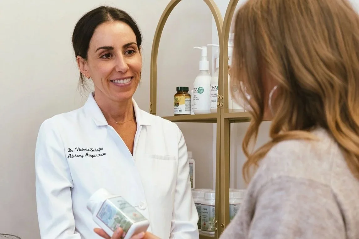 A smiling woman in a white coat with the embroidered name "Dr. Victoria Schefer" and the title "Alchemy Acupuncture" on her chest, holding a product, while speaking with a patient in a medical office.
